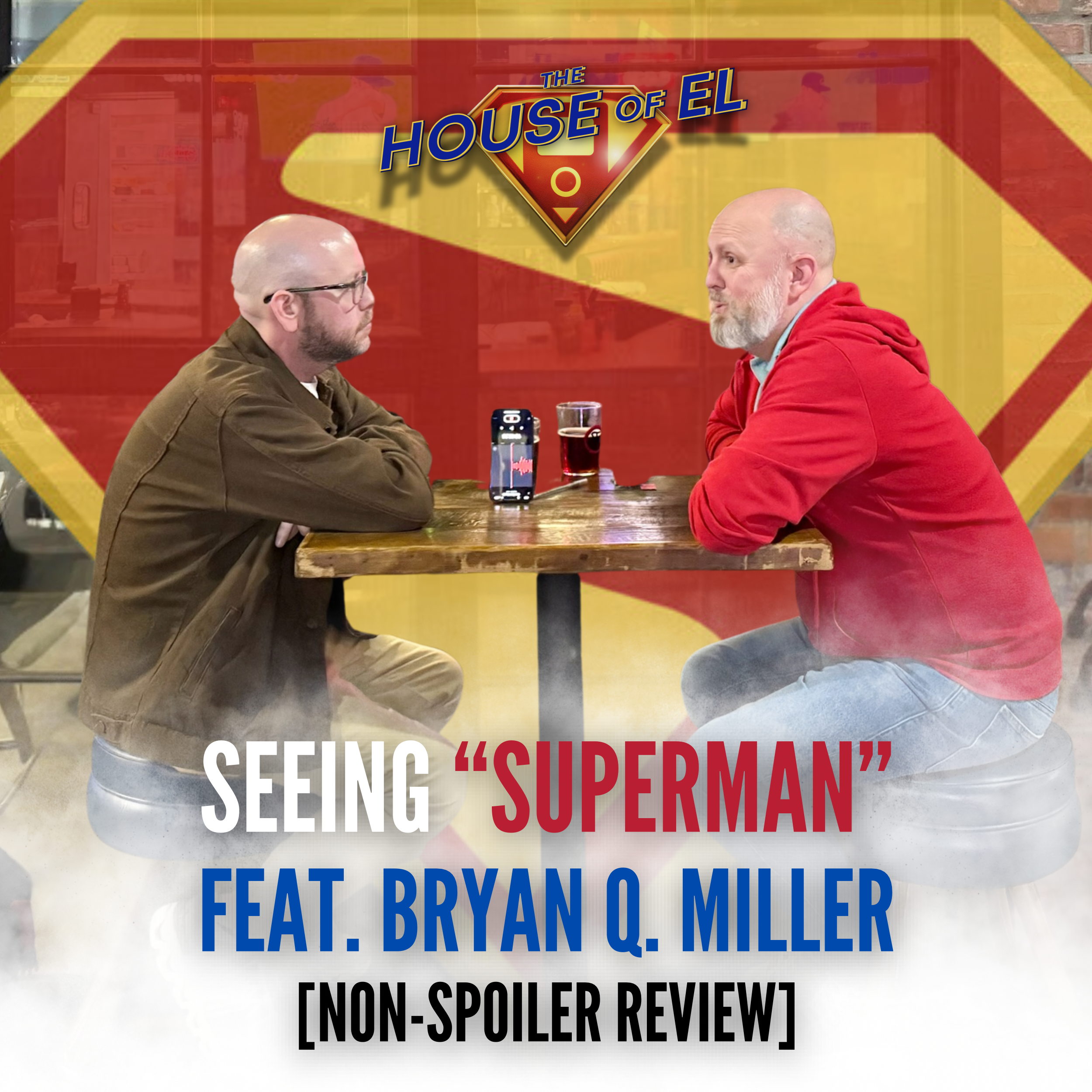 Two men sit at a table in front of a Superman logo. 