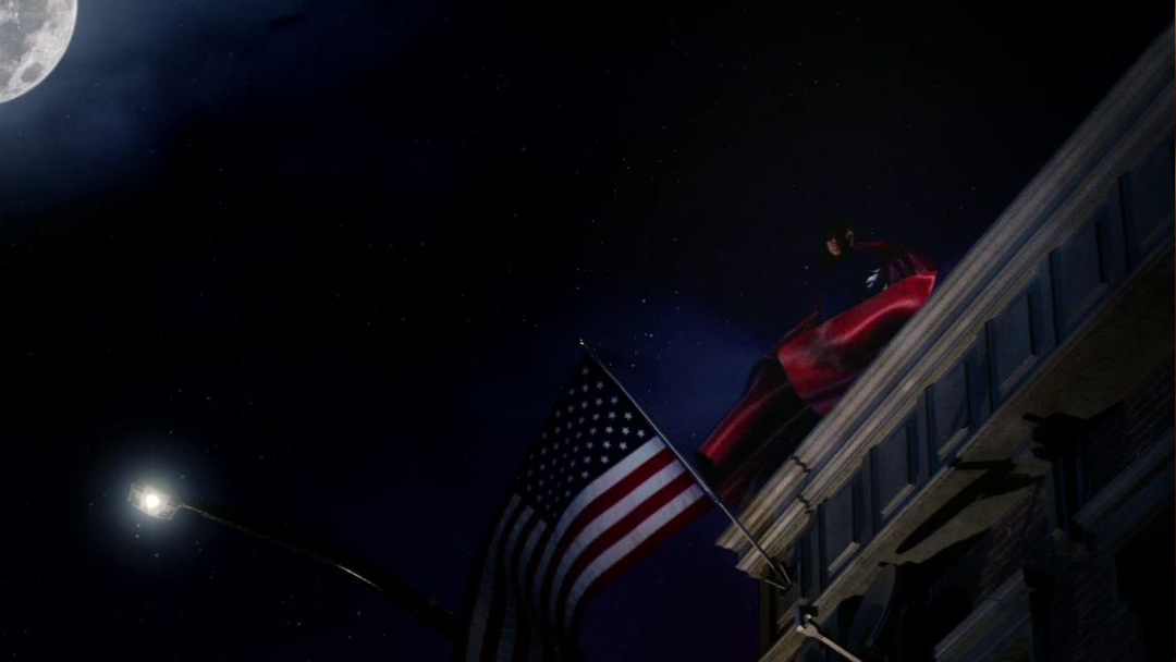 A man in a blue leather jacket and a red cape stands atop a building.