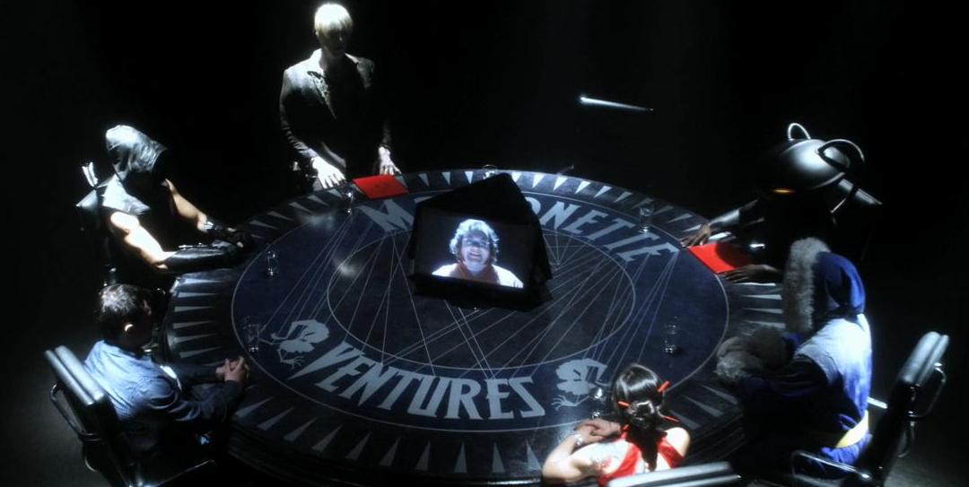 Toyman on a monitor on a table at which Metallo, Roulette, Captain Cold, Black Manta, Solomon Grundy, and the Dark Archer are sitting.