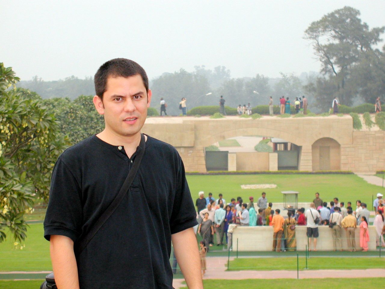 Un viajero posa frente a un jardín donde un grupo de visitantes rodea un monumento.