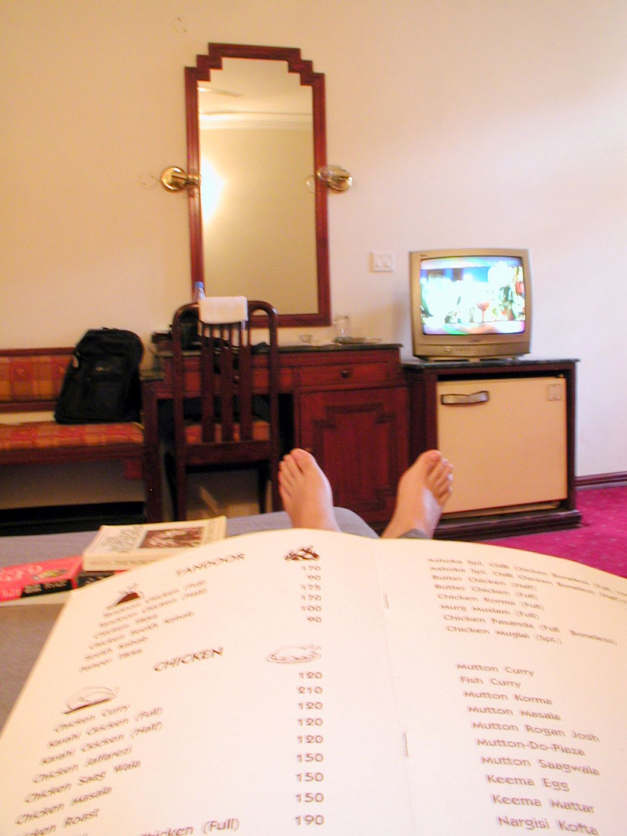 Vista desde una cama de hotel: un menú abierto, un escritorio con espejo y un televisor encendido.