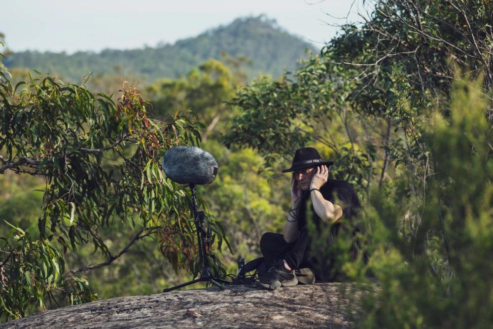 An picture of Lawrence English taken by Greg Harms. It shows Lawrence in a forest with recording equipment.