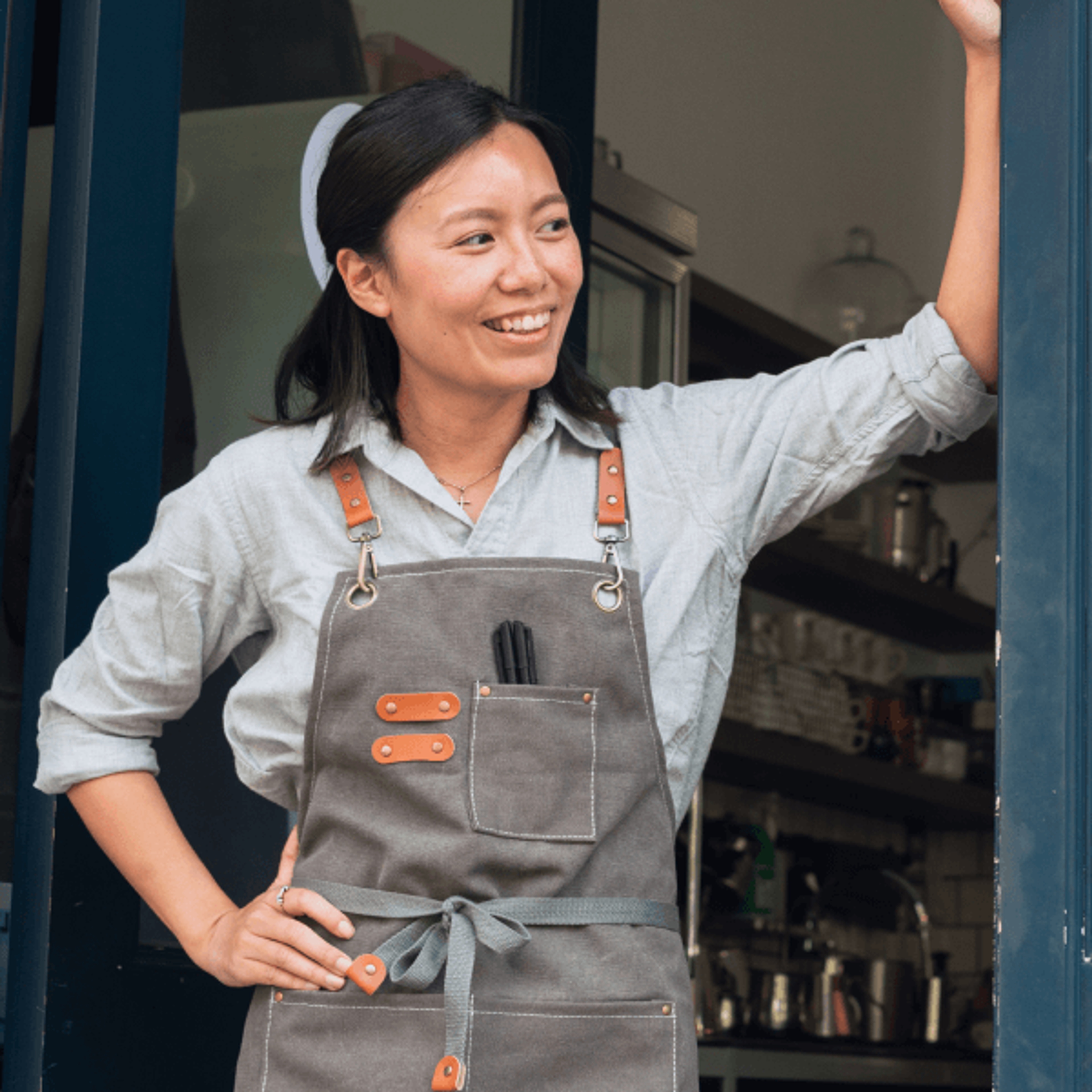 smiling woman in a gray apron with leather straps, standing in a doorway.