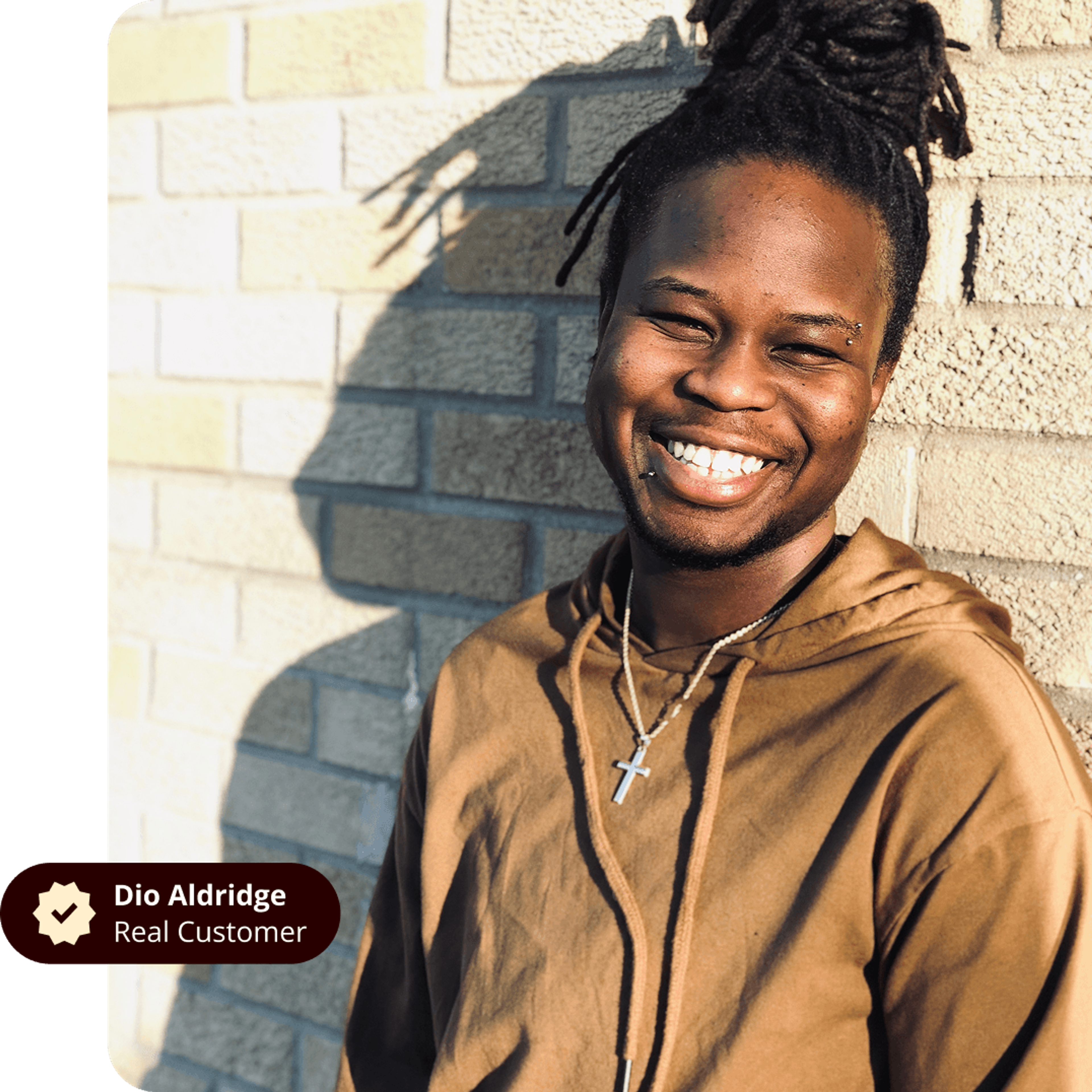 Dio Aldridge, a smiling Black man with dreadlocks and lip piercing, wearing a brown hoodie and cross necklace, stands against a brick wall.