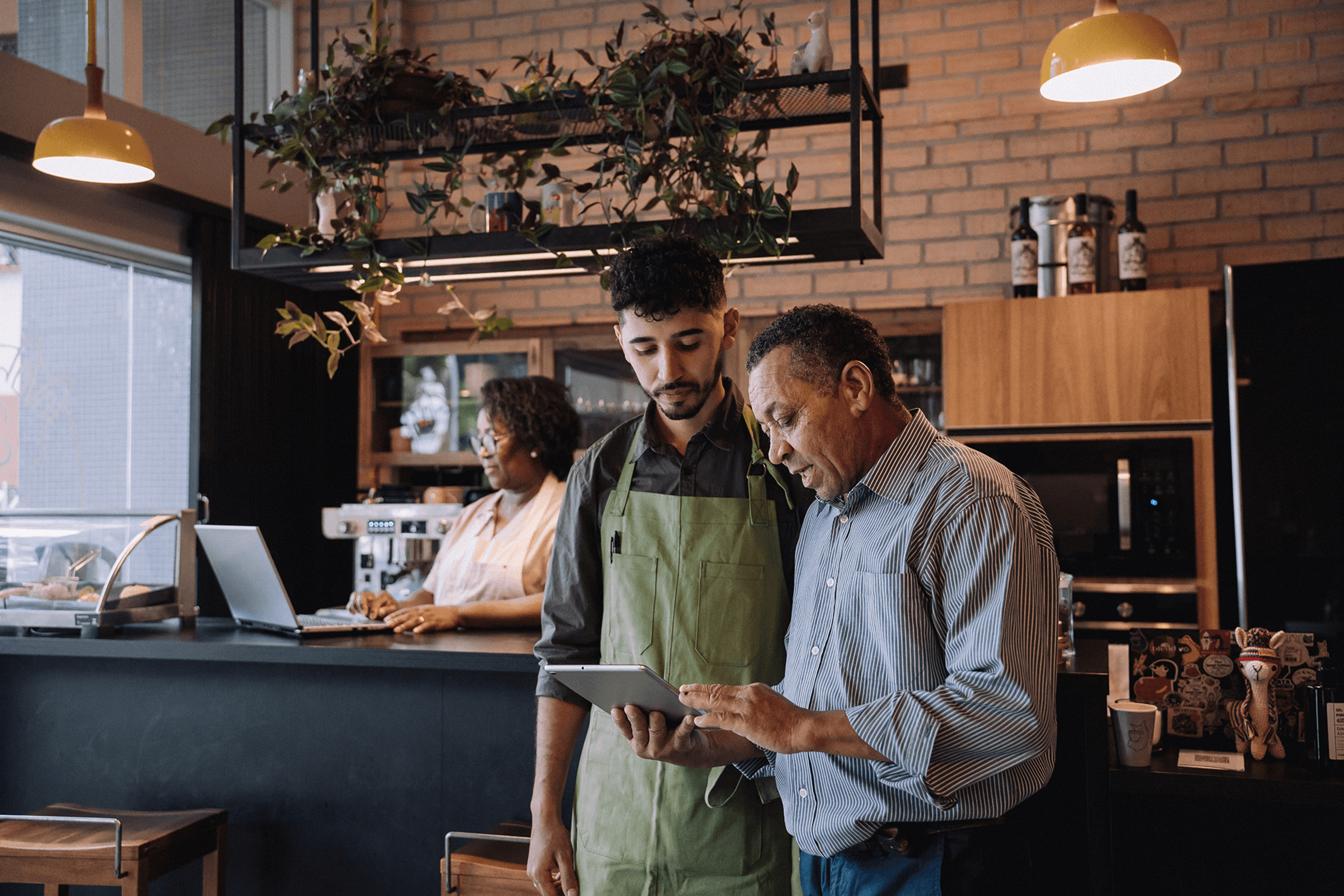 A barista shows a tablet to an older man in a modern cafe, with a woman working on a laptop in the background.