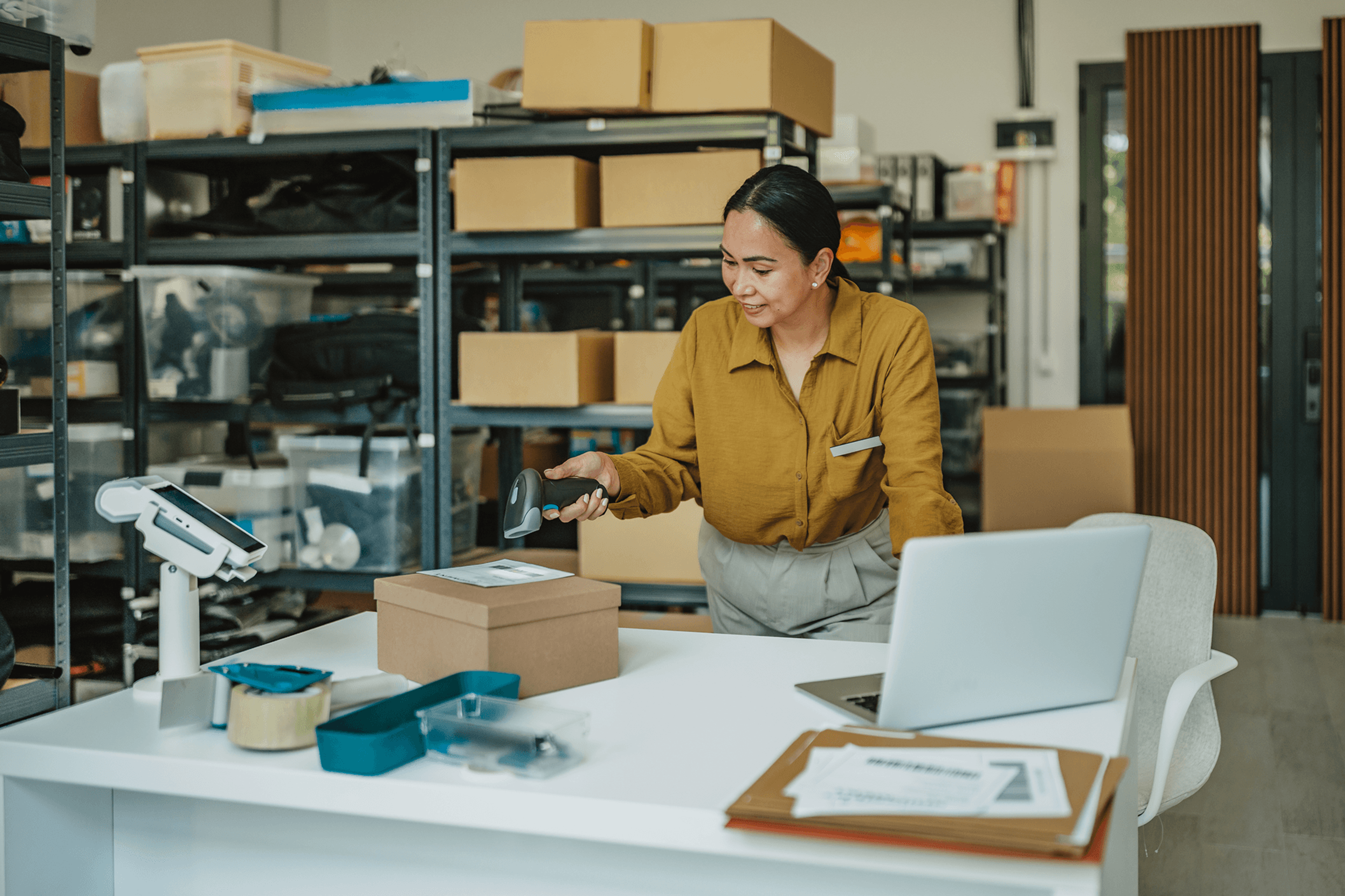 A woman scanning a package on a desk in a storage room.
