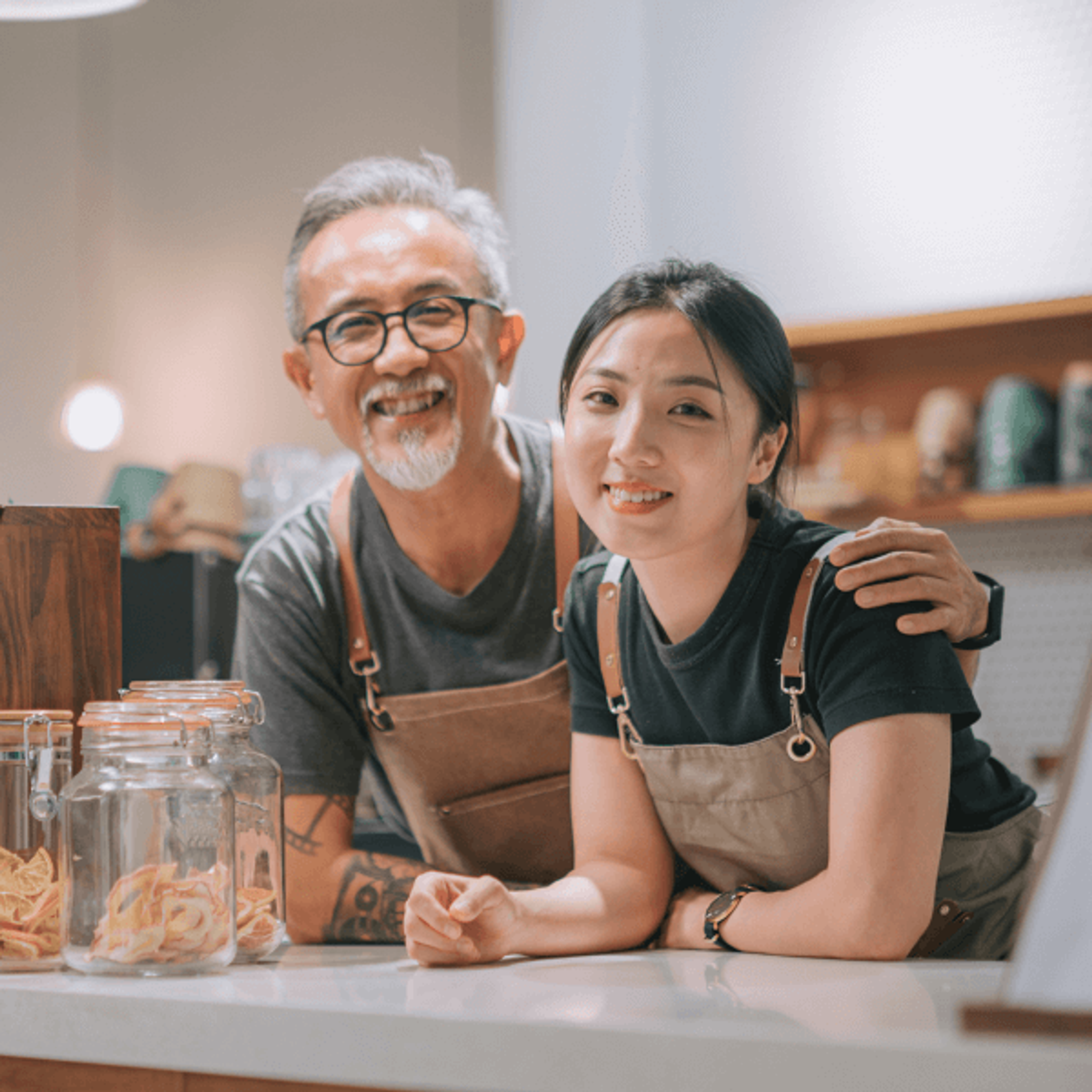 two smiling small business owners, an older man and a younger woman in aprons, stand behind a counter.