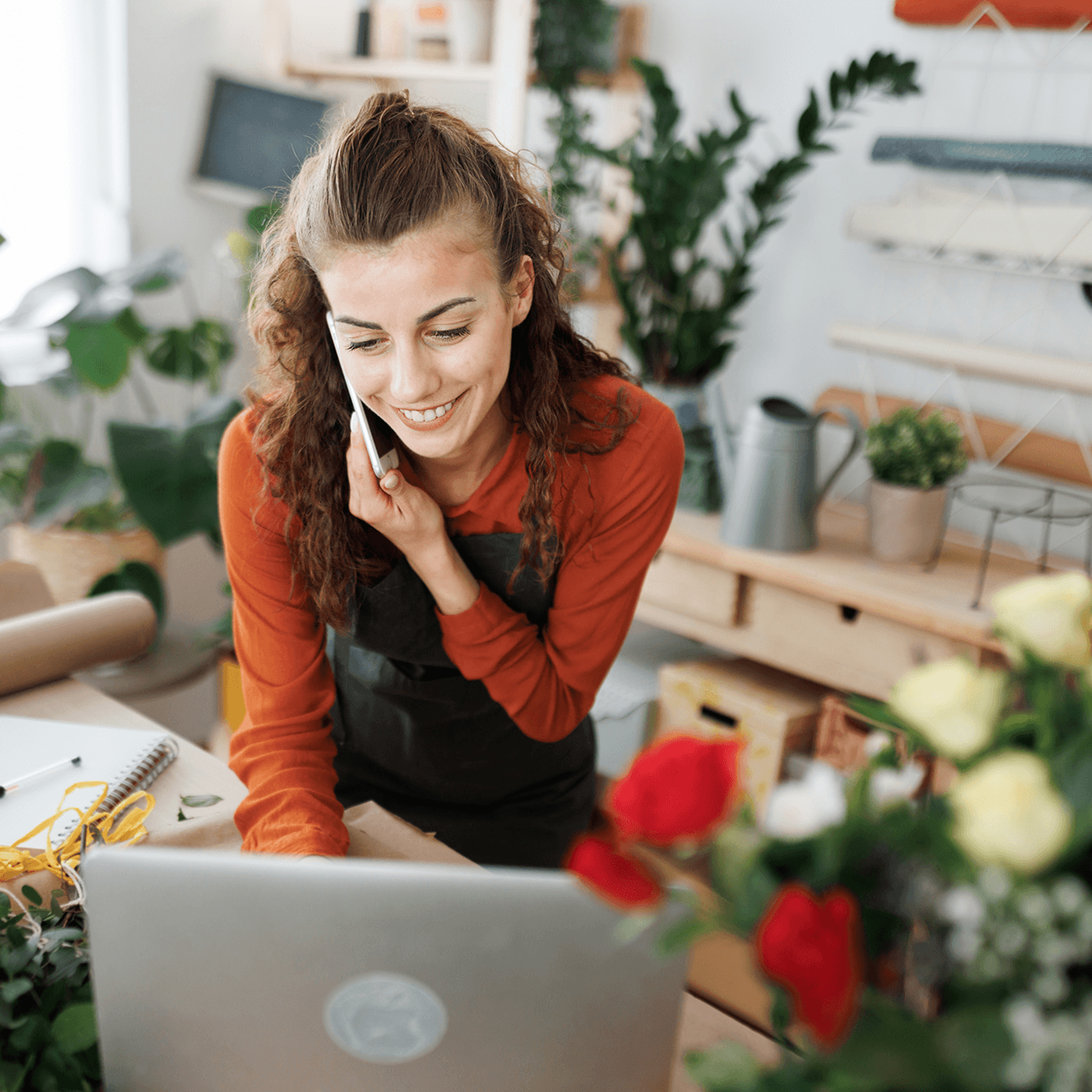 Smiling woman on phone, using laptop in a flower shop.