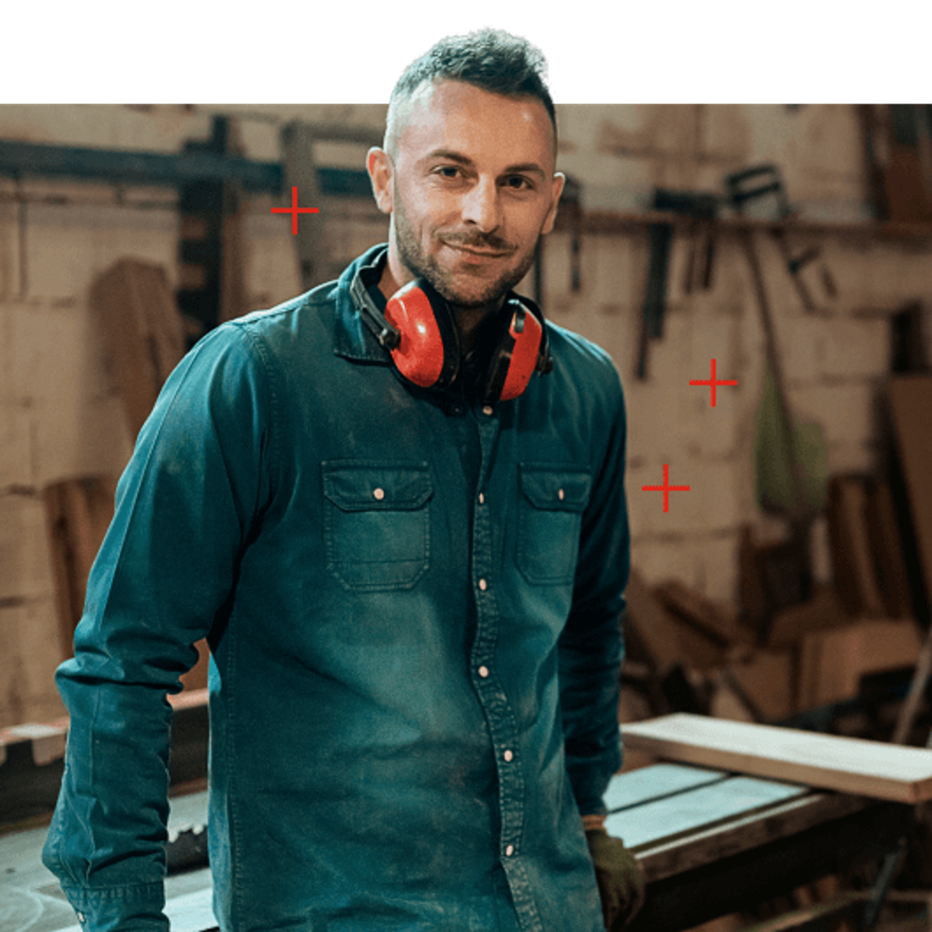 A smiling man in a workshop with red earmuffs around his neck.