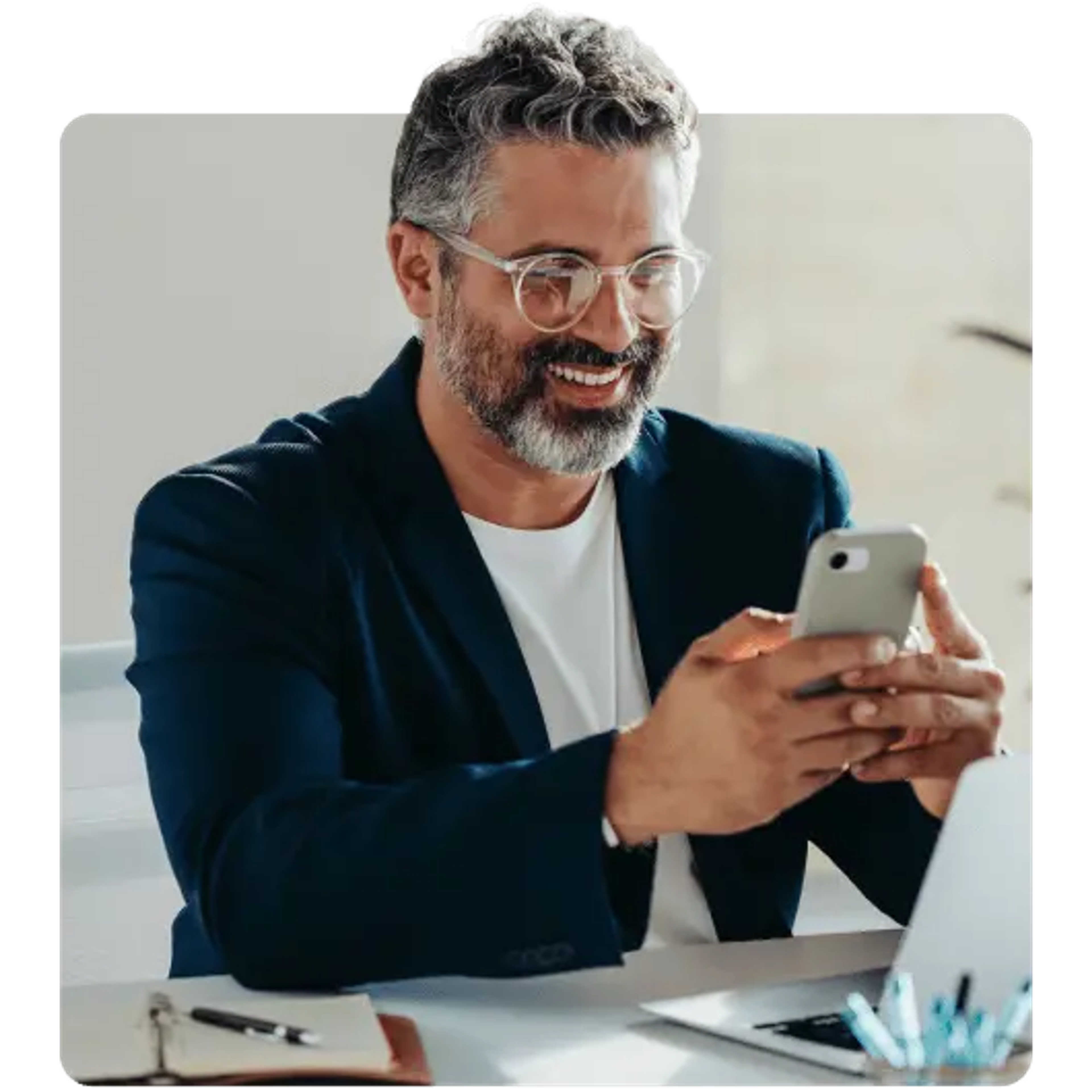 smiling man with glasses and a beard using a smartphone at a desk.