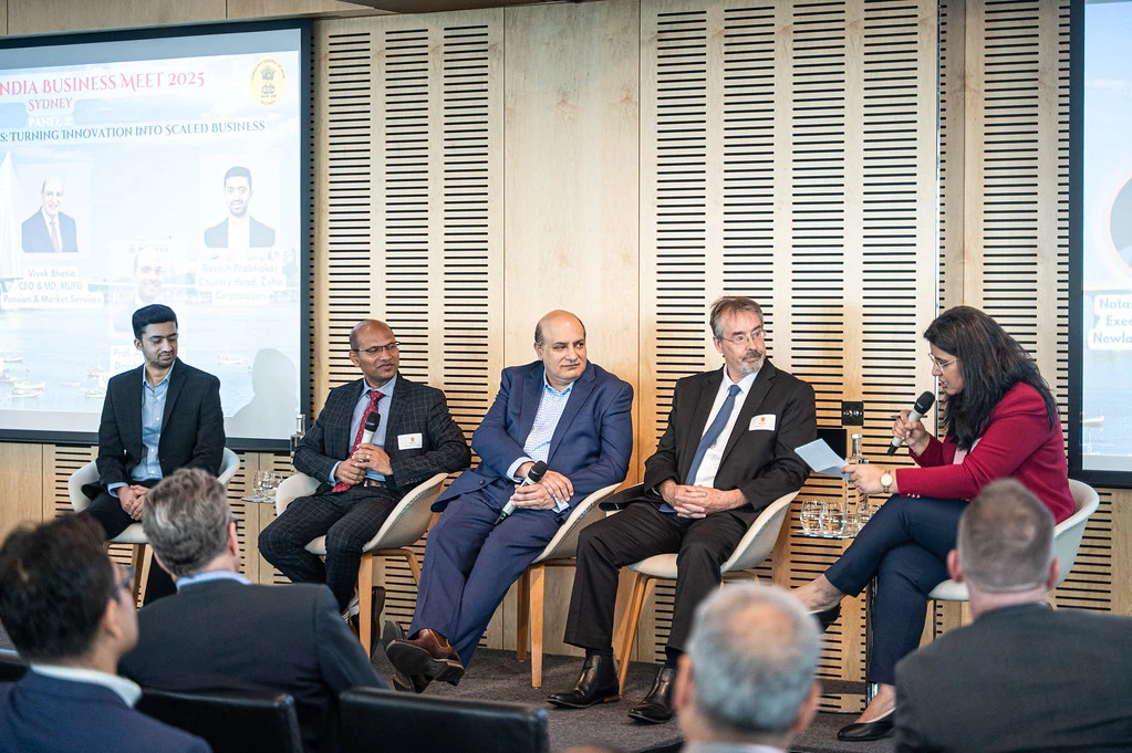 A panel of five speakers sits on stage discussing at the Australia–India Business Meet 2025, with Natasha Jha Bhaskar as an interviewer holding a microphone.