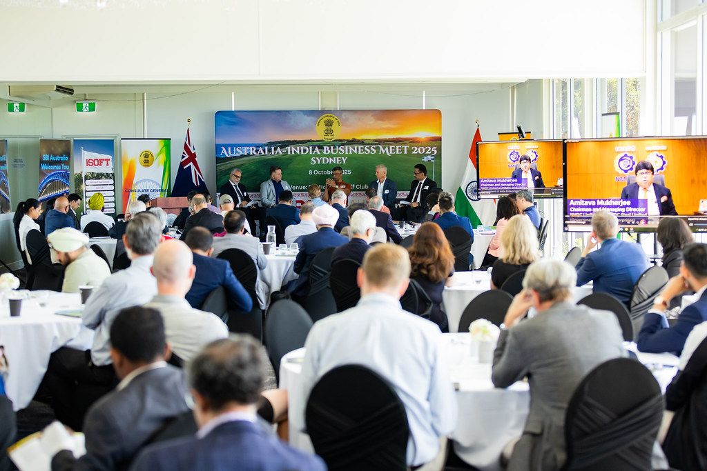 Audience members sit at round tables listening to a panel at the Australia–India Business Meet 2025, with large screens and banners behind the speakers.