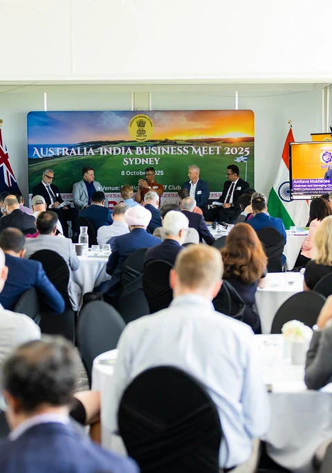 Audience members sit at round tables listening to a panel at the Australia–India Business Meet 2025, with large screens and banners behind the speakers.