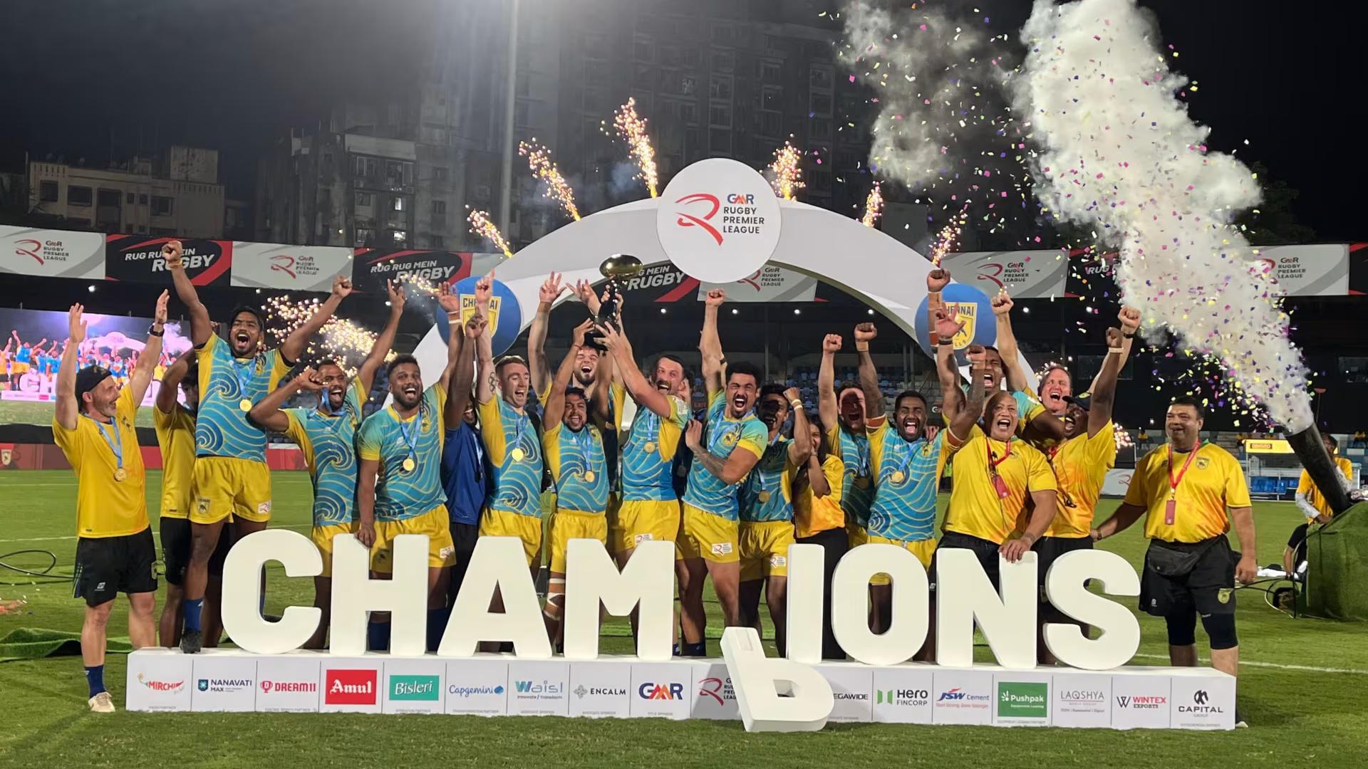 The Chennai rugby team celebrates on the field, lifting the championship trophy with fireworks and confetti behind them, standing in front of large “CHAMPIONS” letters.