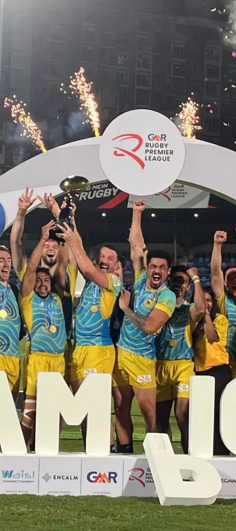 The Chennai rugby team celebrates on the field, lifting the championship trophy with fireworks and confetti behind them, standing in front of large “CHAMPIONS” letters.