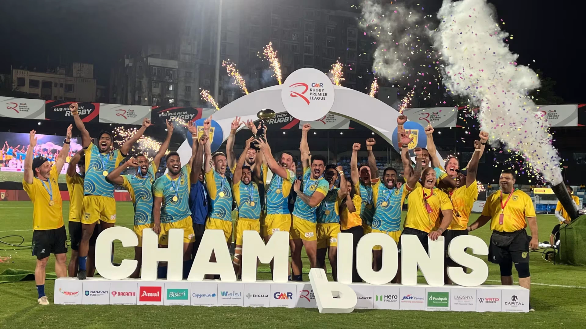 The Chennai rugby team celebrates on the field, lifting the championship trophy with fireworks and confetti behind them, standing in front of large “CHAMPIONS” letters.