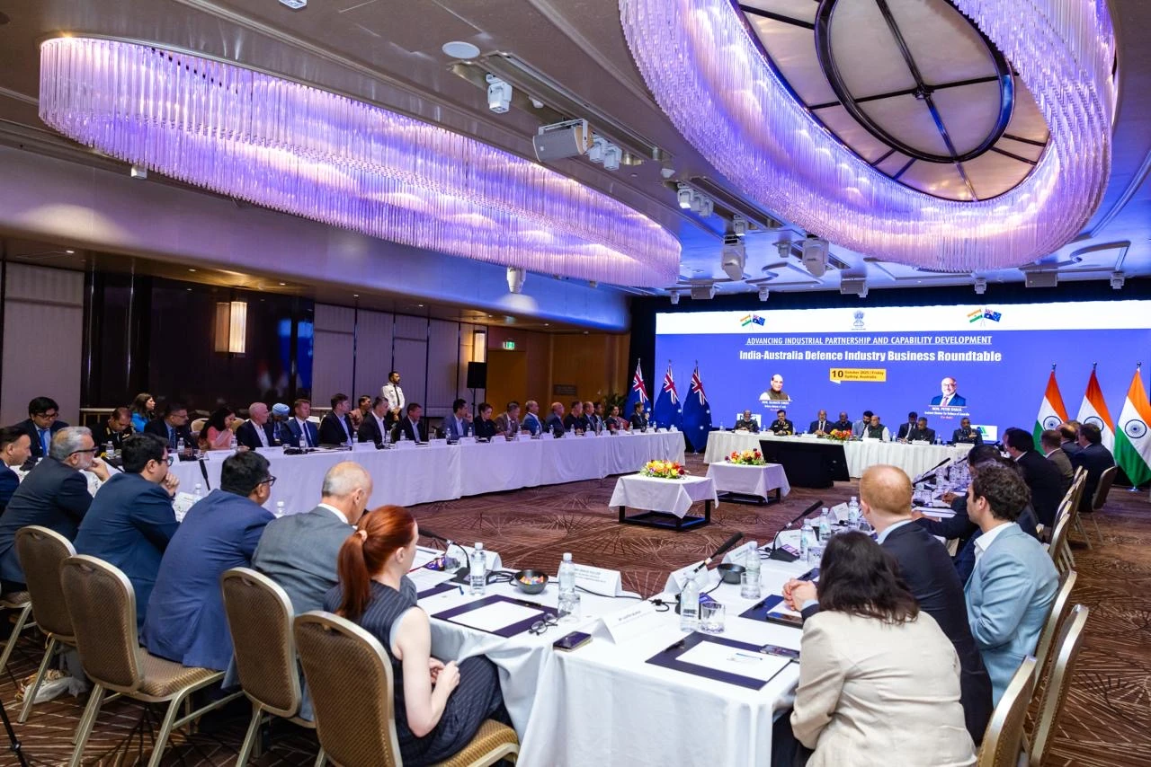 A conference room with Indian and Australian delegates seated around a U-shaped table during the India–Australia Defence Industry Business Roundtable, with flags and a large presentation screen at the front.