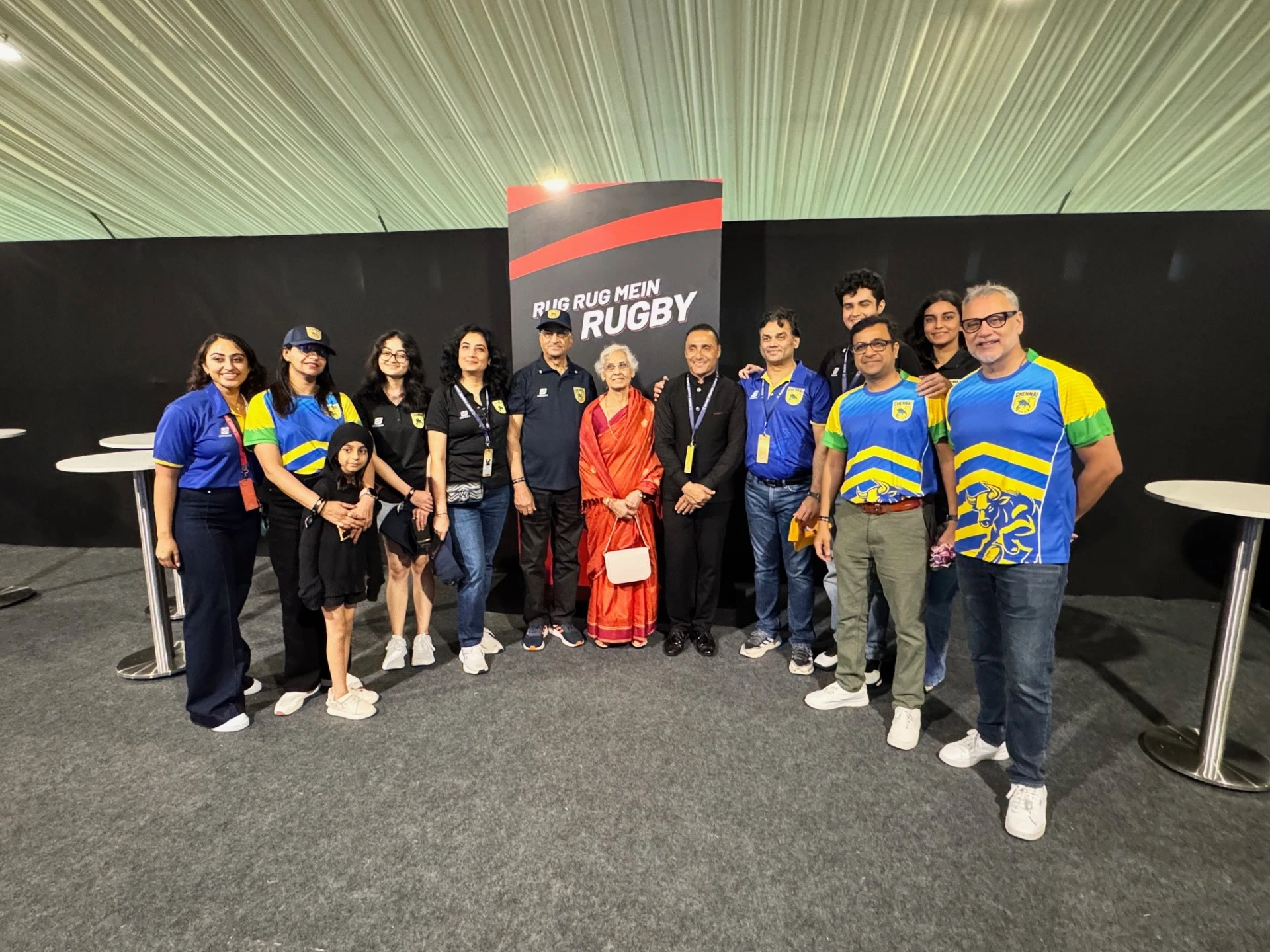 A group of people wearing team and event shirts pose together indoors in front of a “Rug Rug Mein Rugby” backdrop.