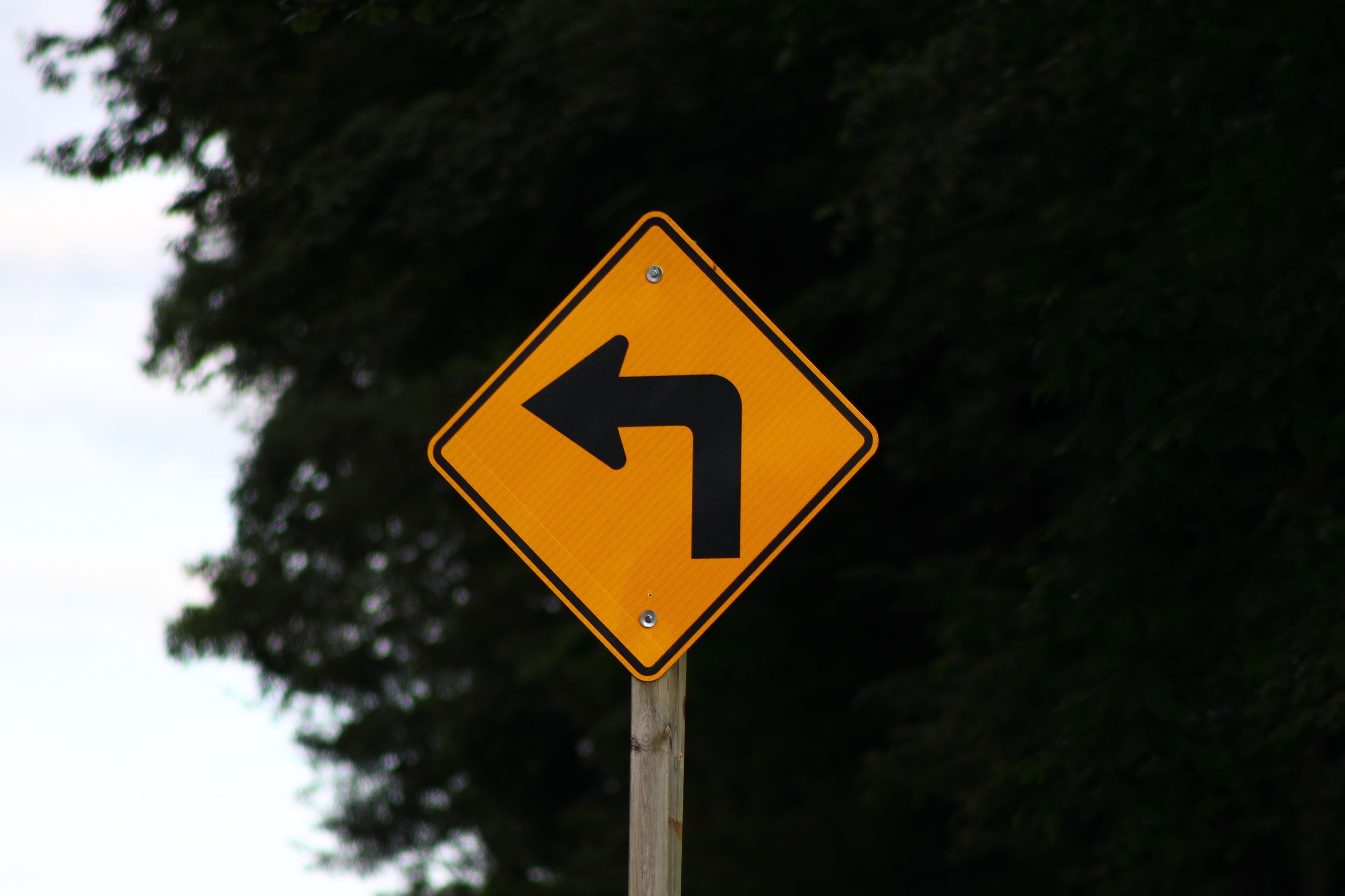 Yellow diamond-shaped left turn warning road sign on a wooden post against a dark tree background, illustrating left-turn accident risks on New Jersey roads