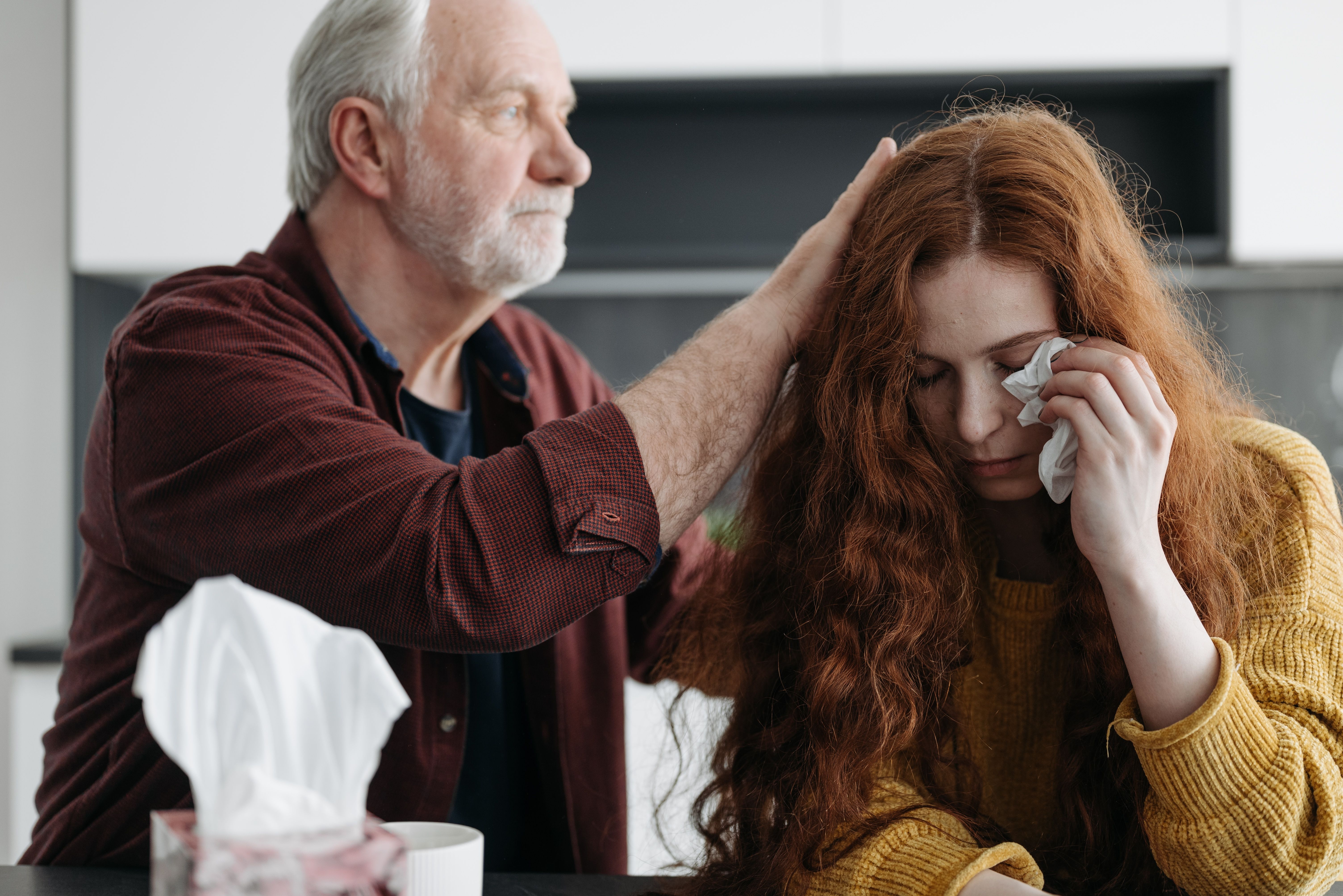 Image Older man comforting a grieving young woman wiping away tears at a kitchen table with a tissue box, after a wrongful death loss in New Jersey