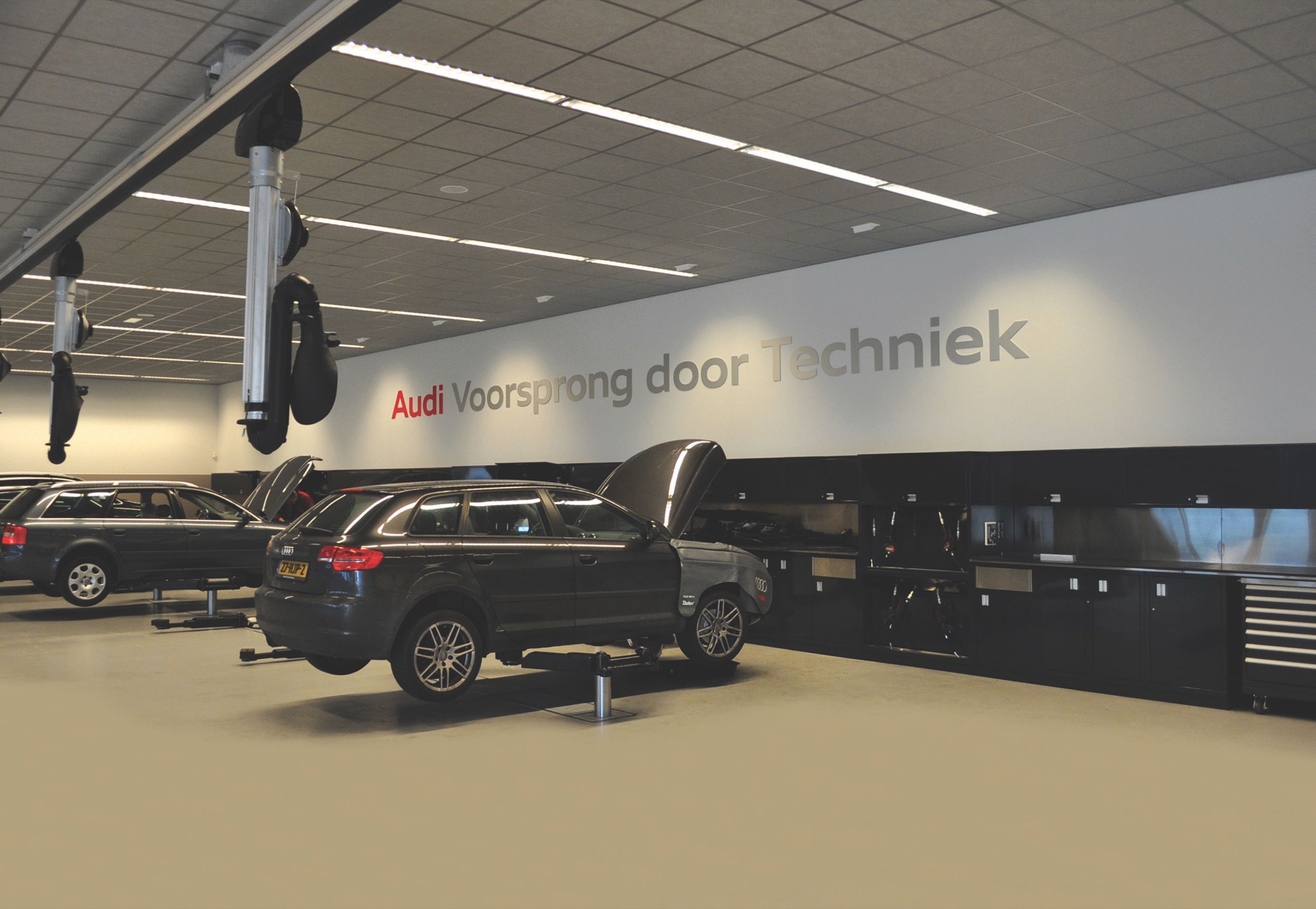Dealership service hall with SUVs on lifts, overhead hose reels and a long run of black workbenches and cabinets