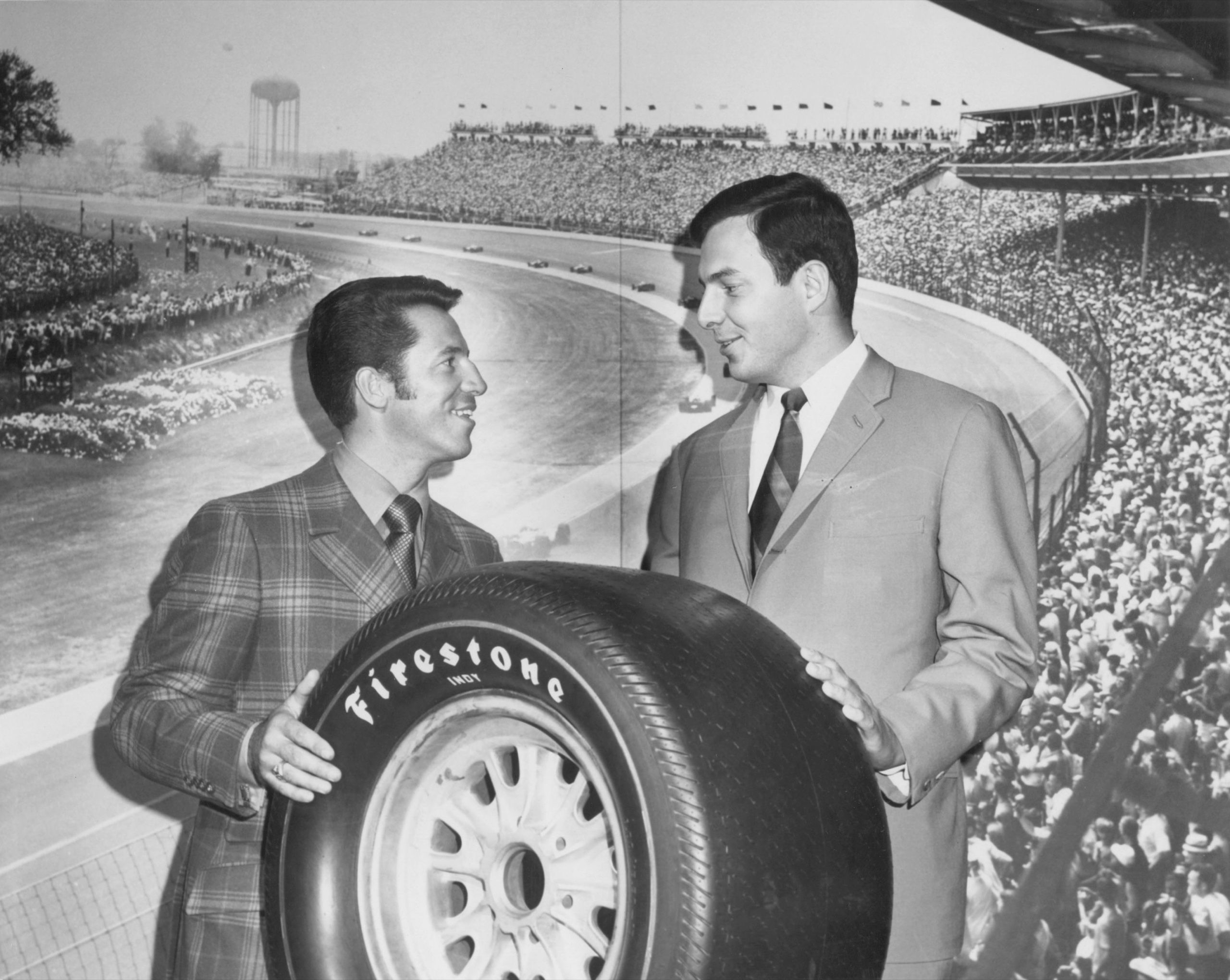 Archival black-and-white photo of two men holding a Firestone tire in front of a racetrack grandstand