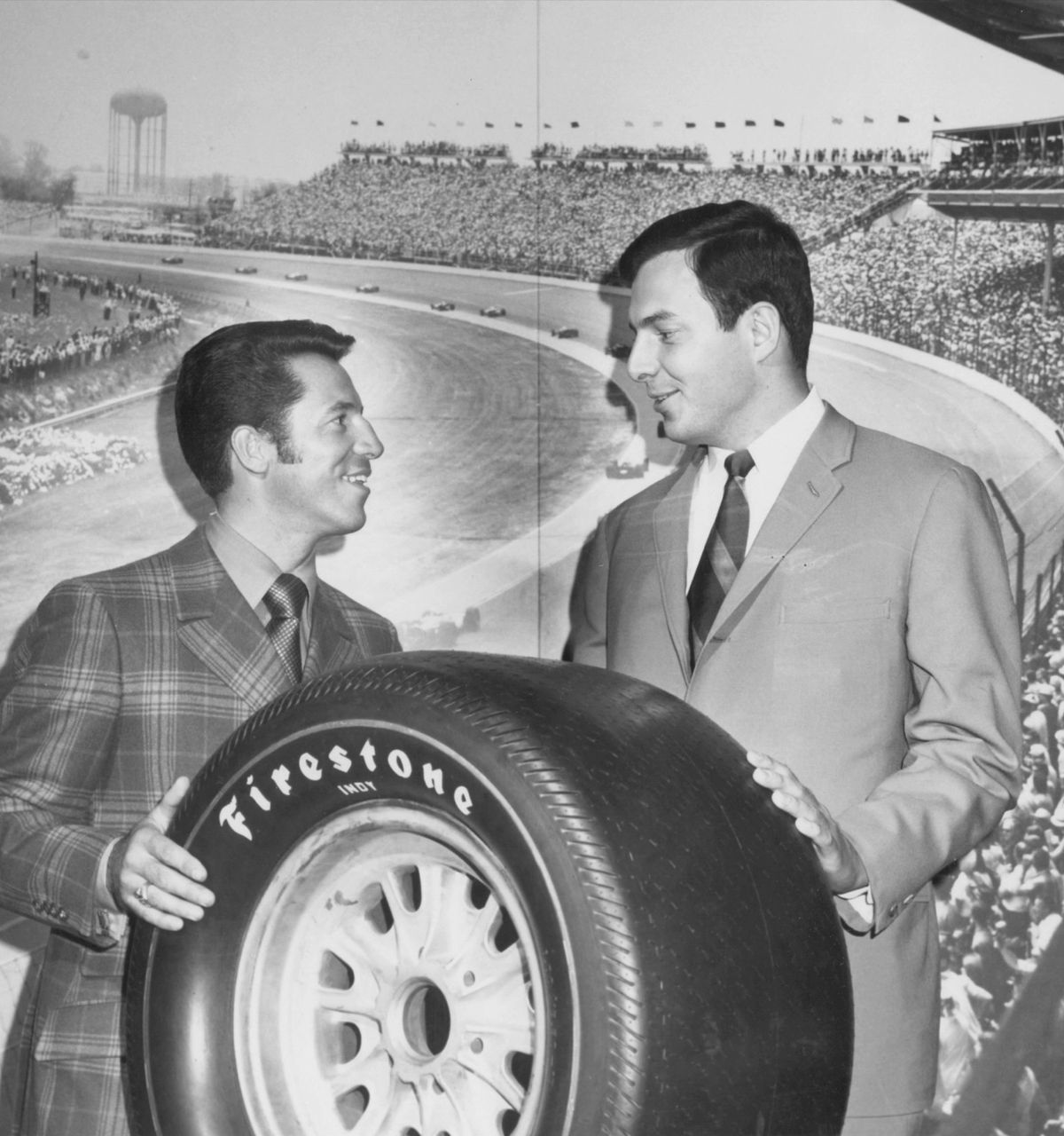 Archival black-and-white photo of two men holding a Firestone tire in front of a racetrack grandstand