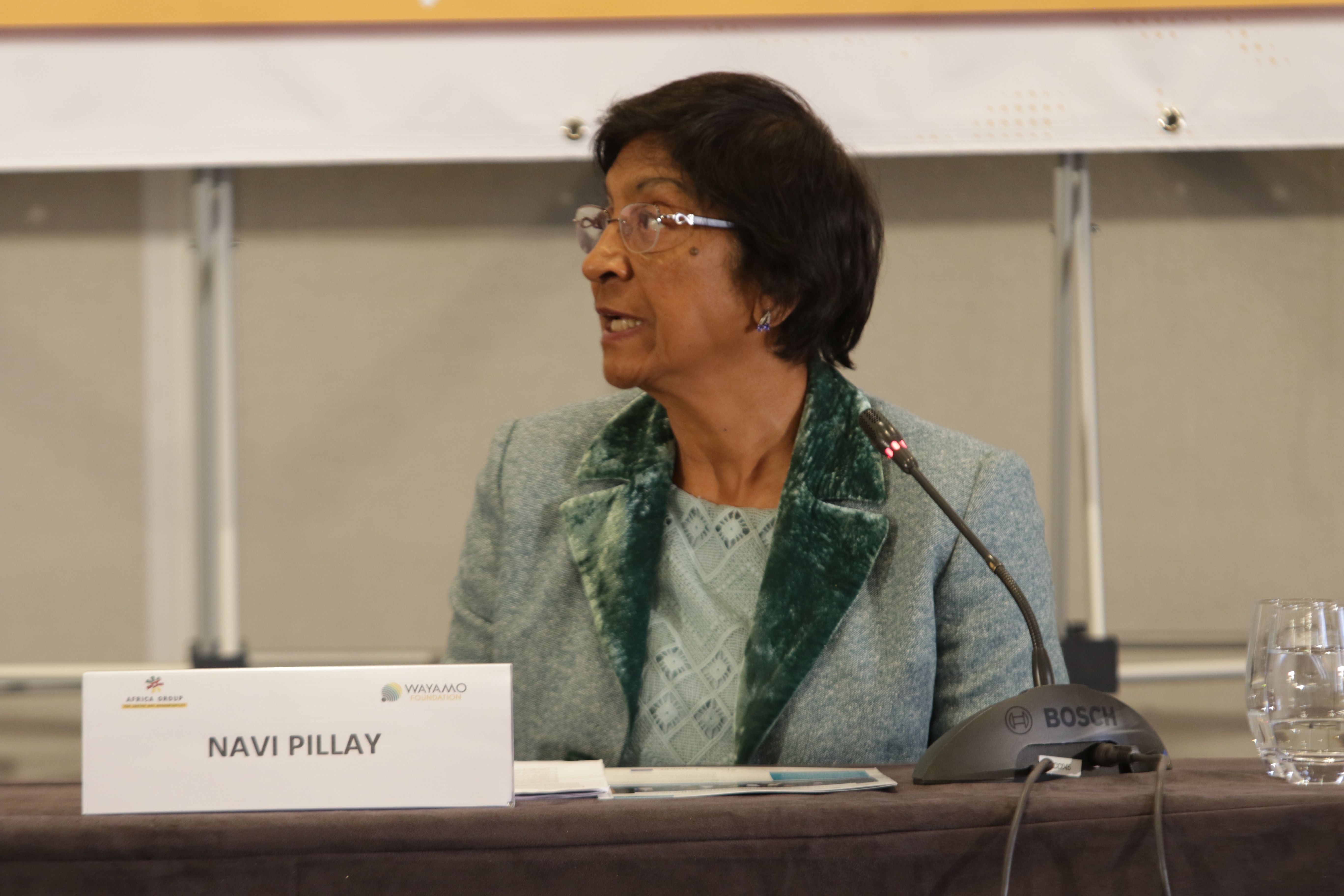 Navi Pillay, con lentes y saco gris con solapa verde, habla sentada frente a un micrófono detrás de una mesa con su gafete de nombre.