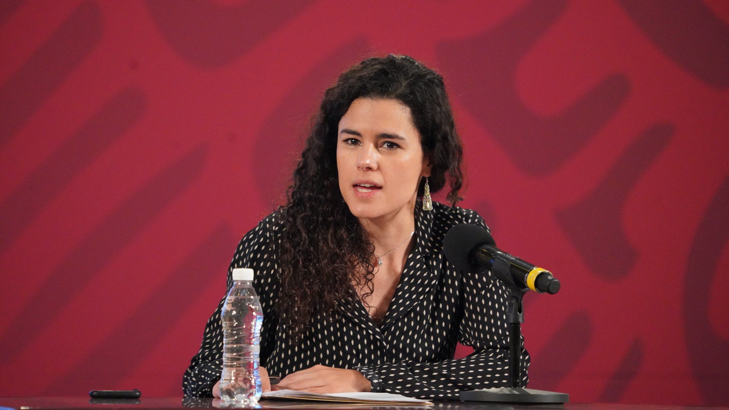 Luisa María Alcalde, de cabello rizado y blusa negra con puntos blancos, habla ante un micrófono sentada a una mesa frente a un fondo rojo con el logotipo del Gobierno de México.