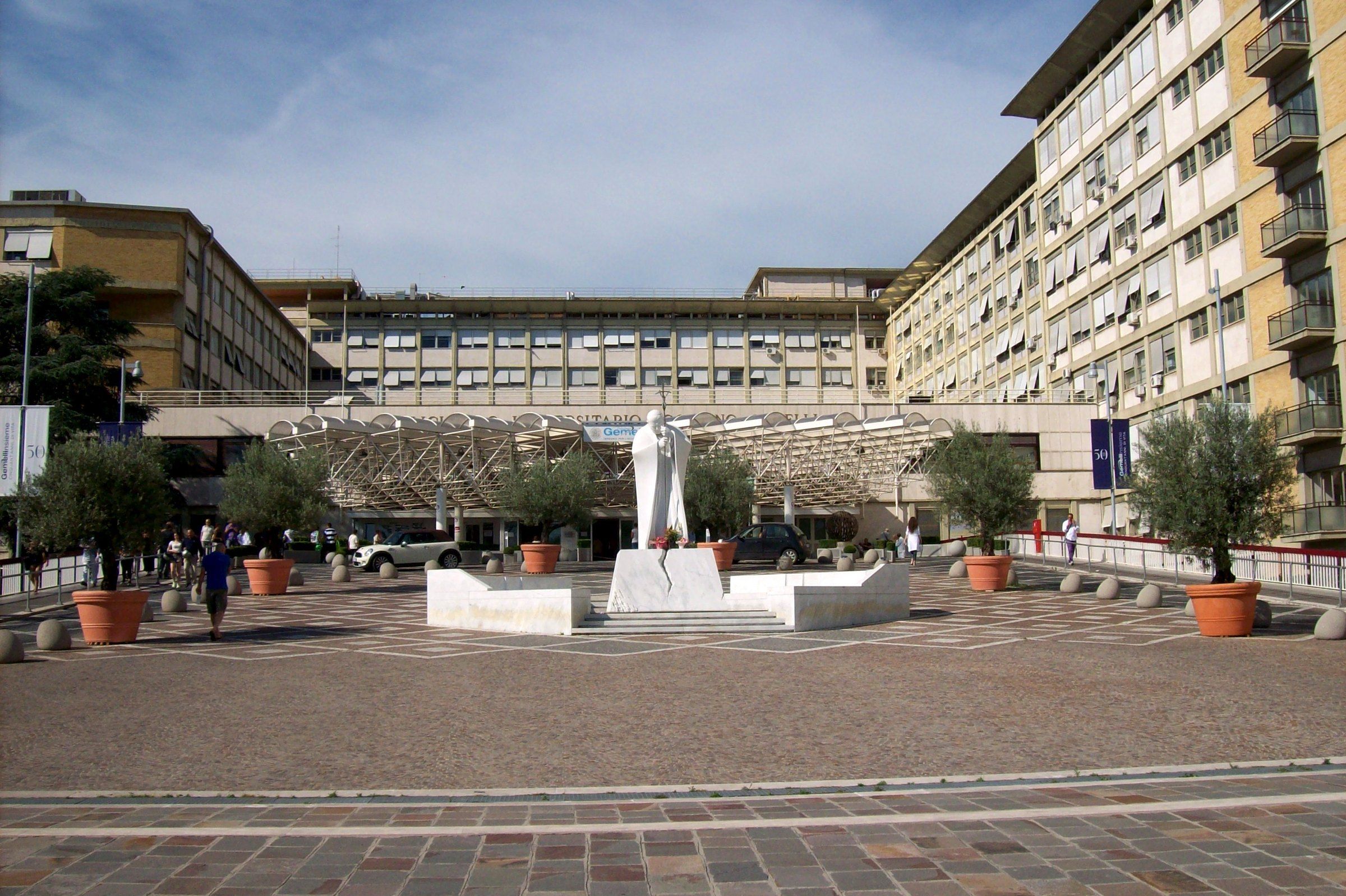 Fachada del Policlínico Agostino Gemelli en Roma, con el edificio hospitalario y árboles en primer plano bajo un cielo claro.