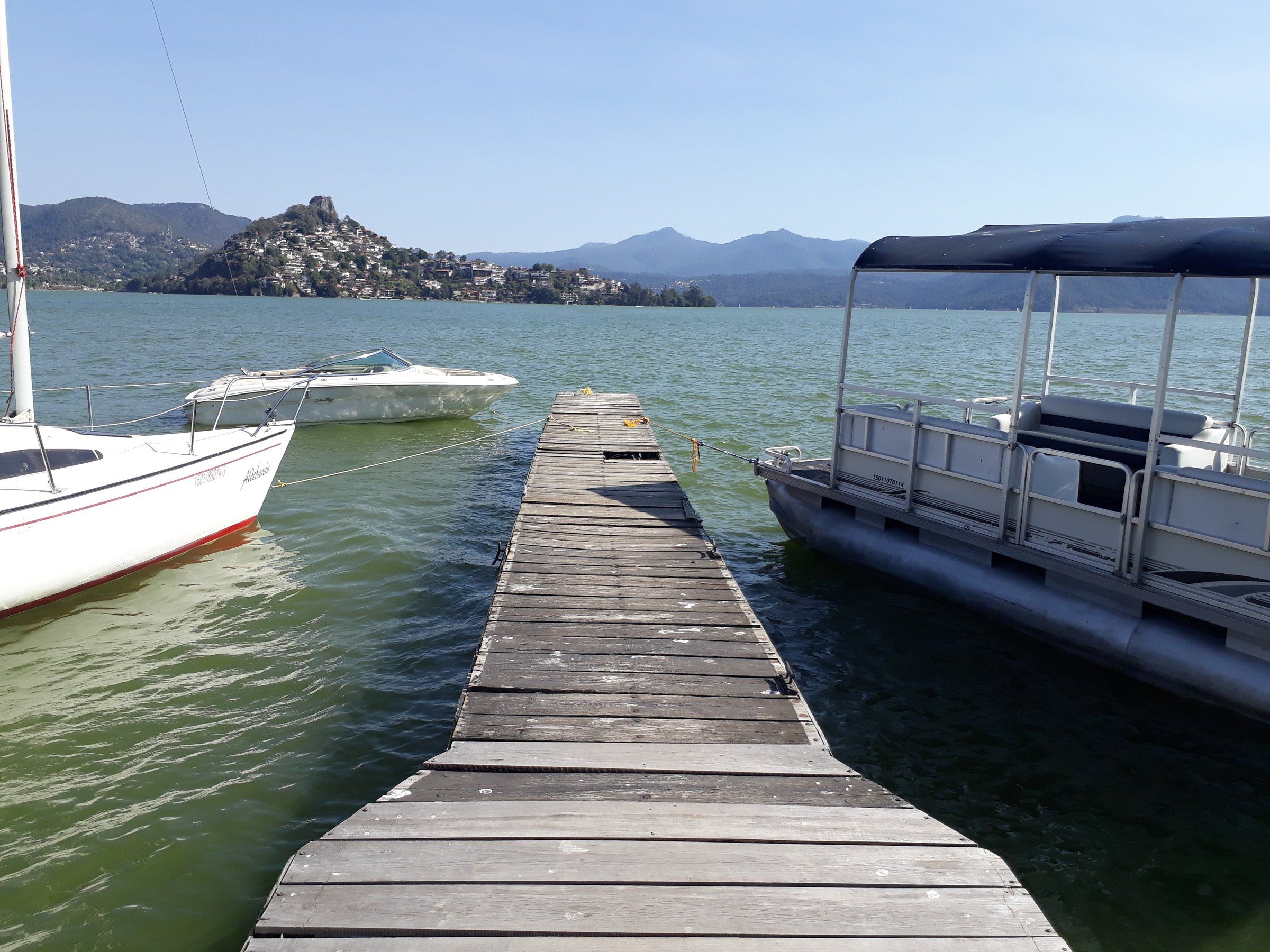 Muelle de madera con lanchas amarradas en el lago de la presa Valle de Bravo, con cerros al fondo bajo un cielo azul.