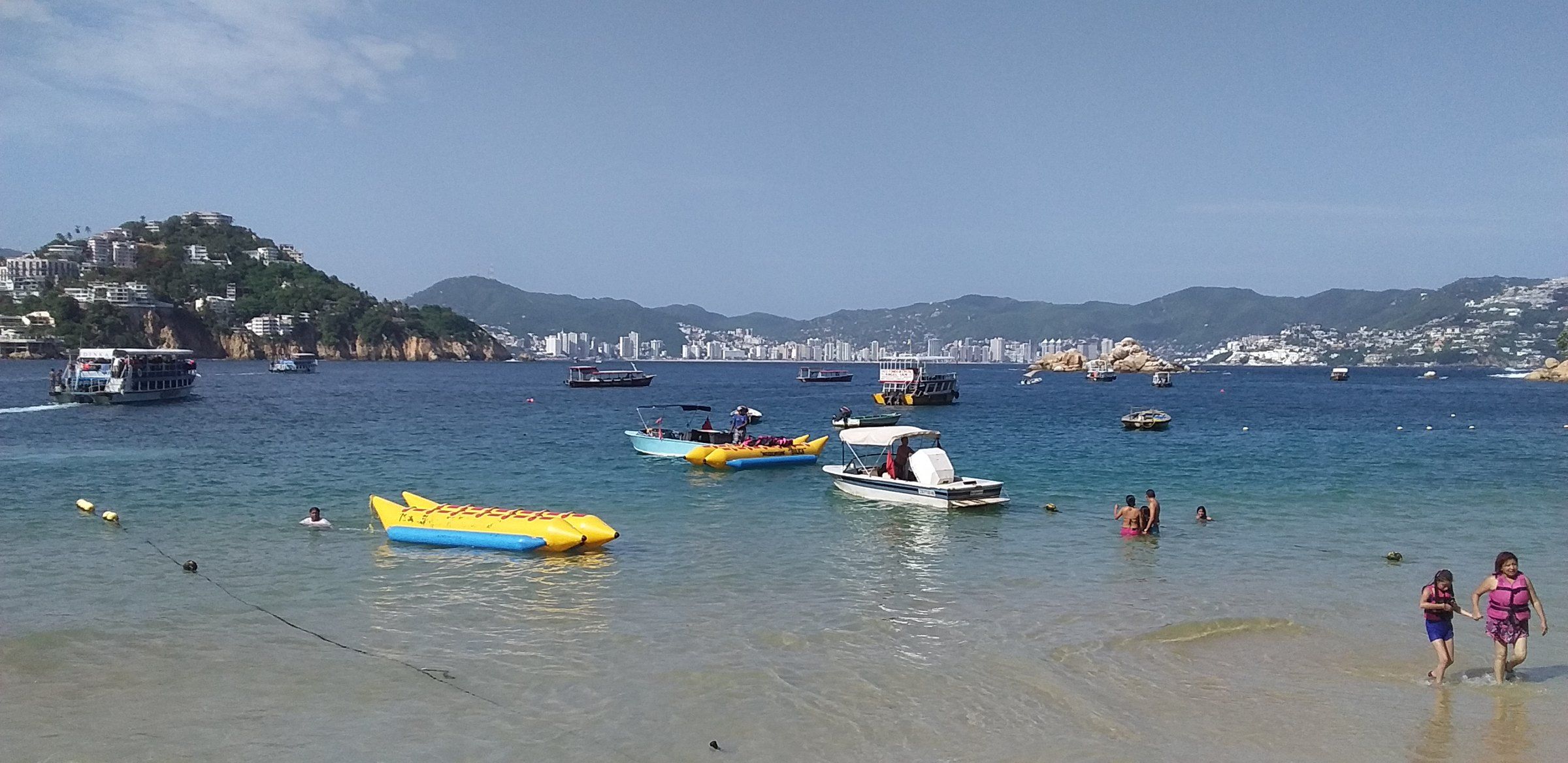 Vista de la bahía de Acapulco con el mar en primer plano, barcos y la franja hotelera al fondo bajo cielo despejado.