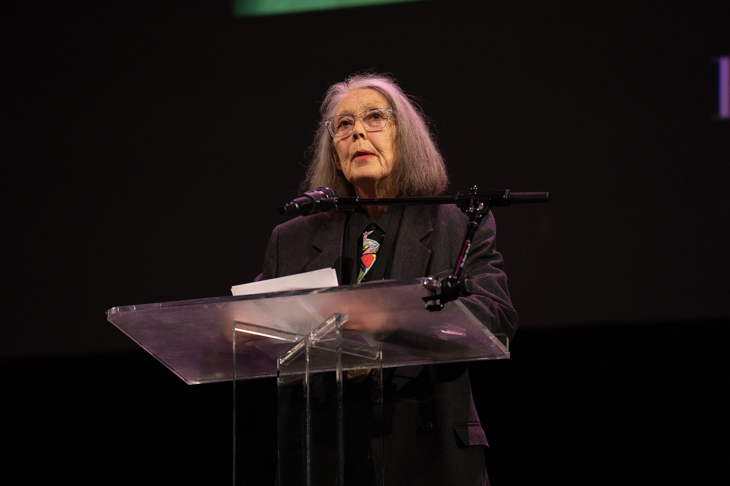 Anne Carson, mujer de cabello canoso y lentes, con saco oscuro y corbata de colores, lee de pie detrás de un atril transparente sobre un escenario con fondo negro.