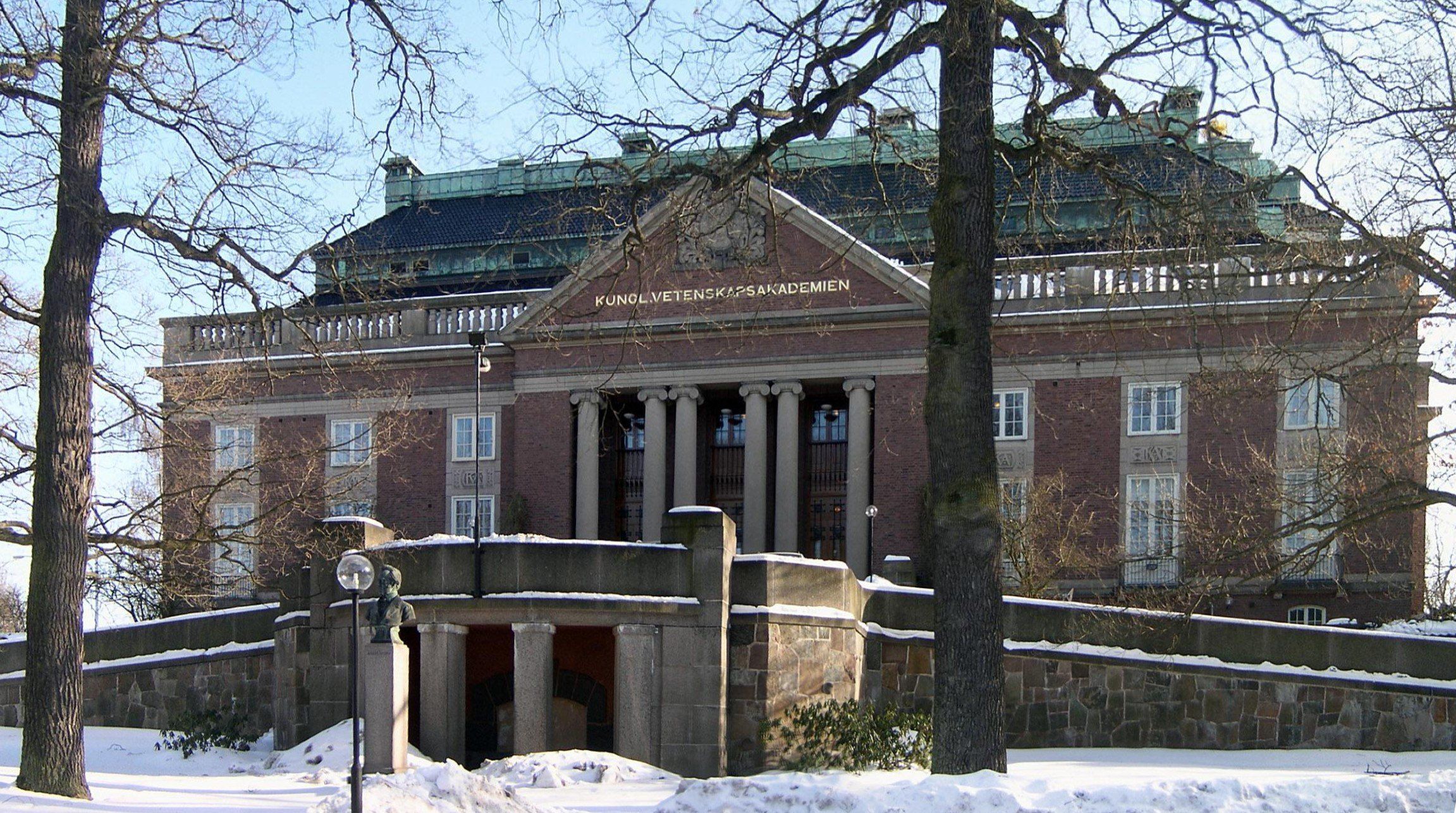Edificio principal de la Real Academia Sueca de Ciencias en Estocolmo, de ladrillo rojo con columnas y la inscripción Kungl. Vetenskapsakademien en el frontón, rodeado de árboles sin hojas y nieve.