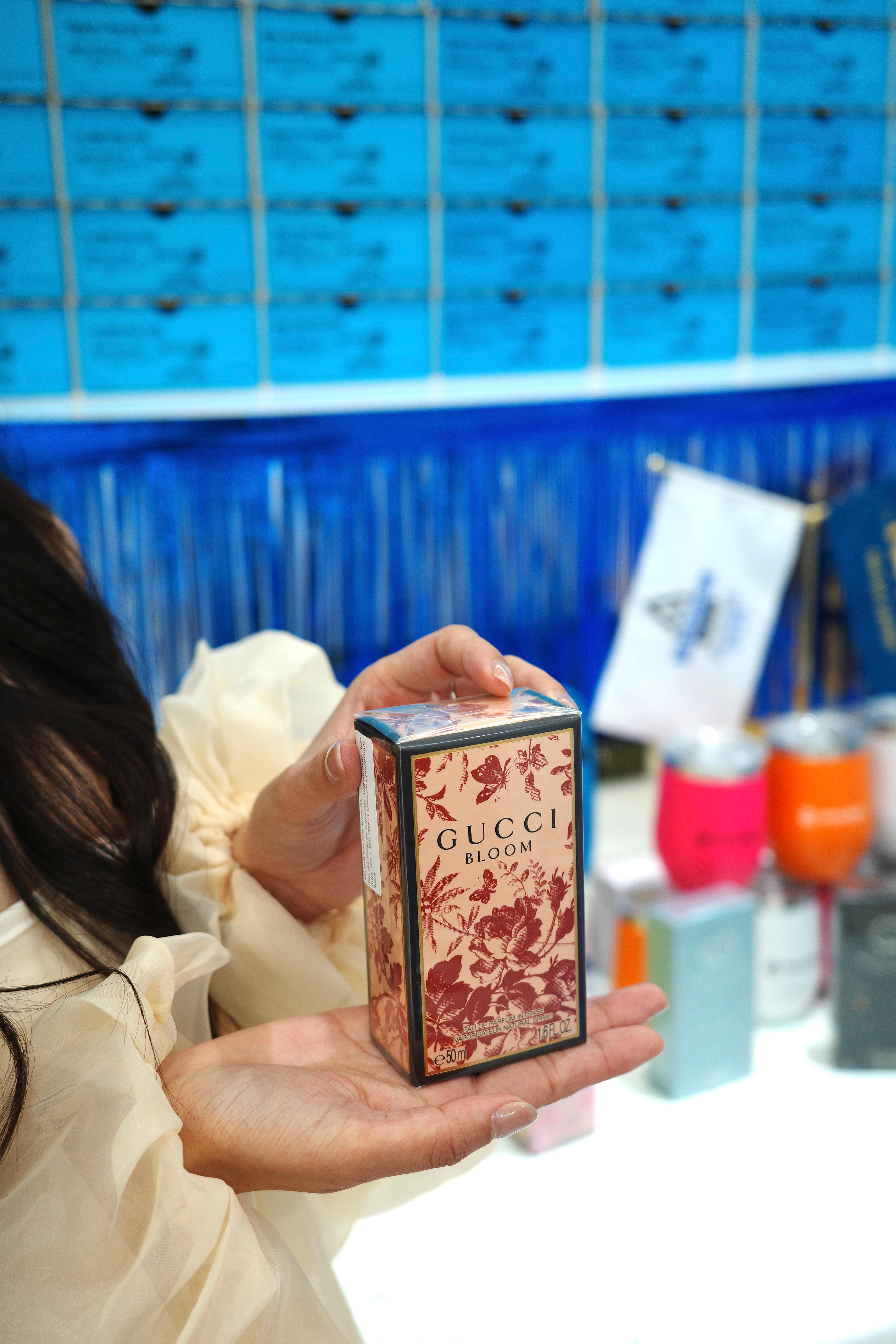 Cận cảnh hai bàn tay cầm hộp nước hoa Gucci Bloom trong phần quà của sự kiện; phía sau là bức tường hộp Invisalign xanh và bàn quà đã bày sẵn.