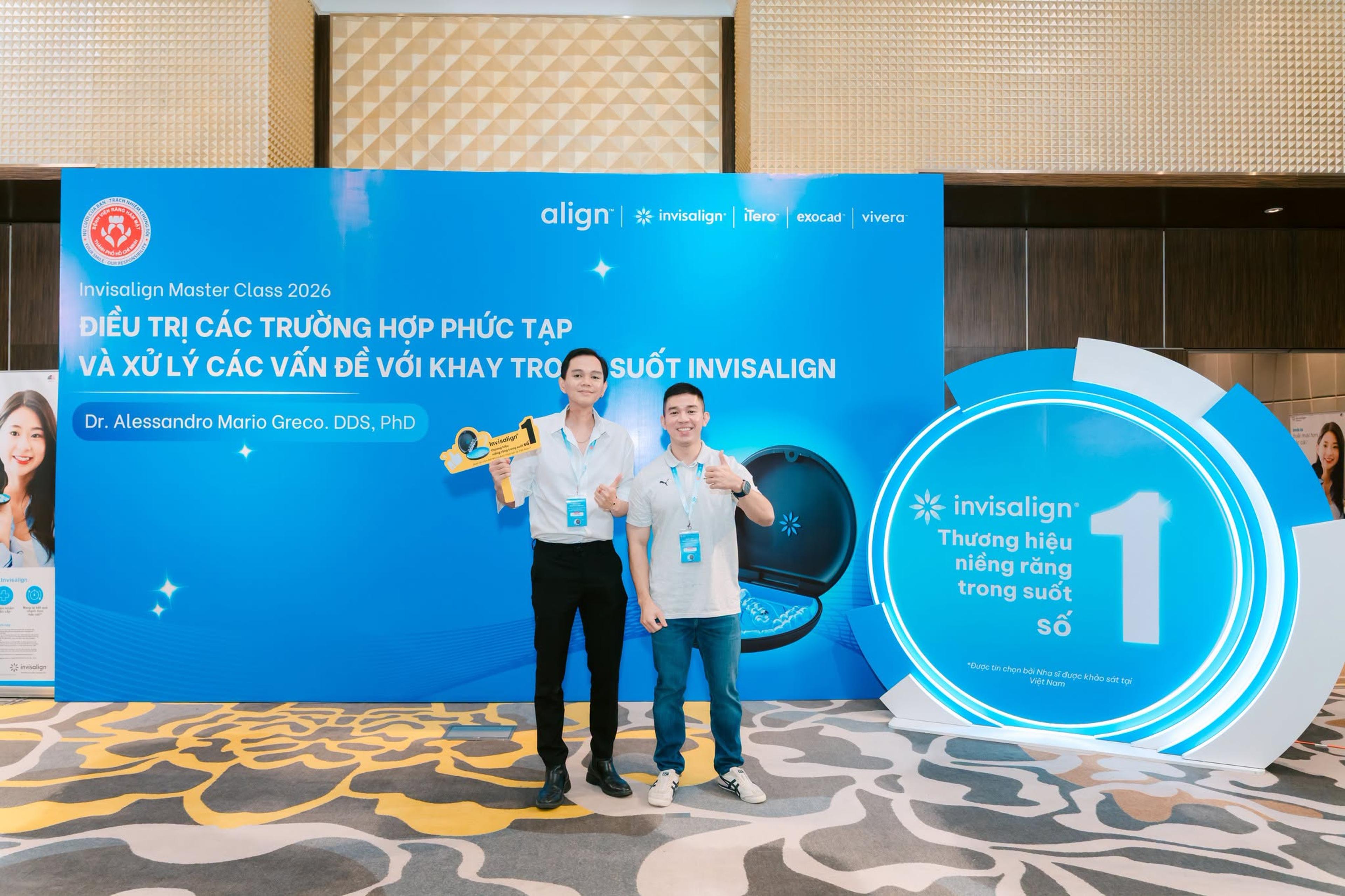 Two Dr Tony Dental clinicians in front of a backdrop reading “Invisalign Master Class 2026 — Treating complex cases and troubleshooting with Invisalign clear aligners, Dr. Alessandro Mario Greco, DDS, PhD”; an Invisalign promotional stand is to the right.
