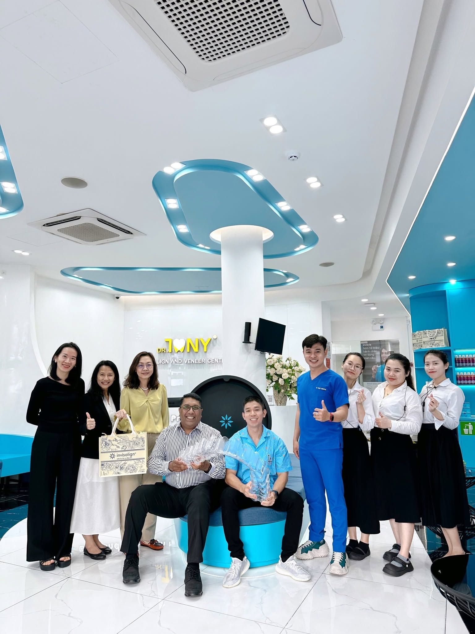 Đoàn lãnh đạo Align Technology chụp ảnh cùng đội ngũ Dr Tony Dental tại sảnh phòng khám; hai khách mời ngồi phía trước, một người cầm túi quà và một người cầm kỷ niệm chương pha lê.