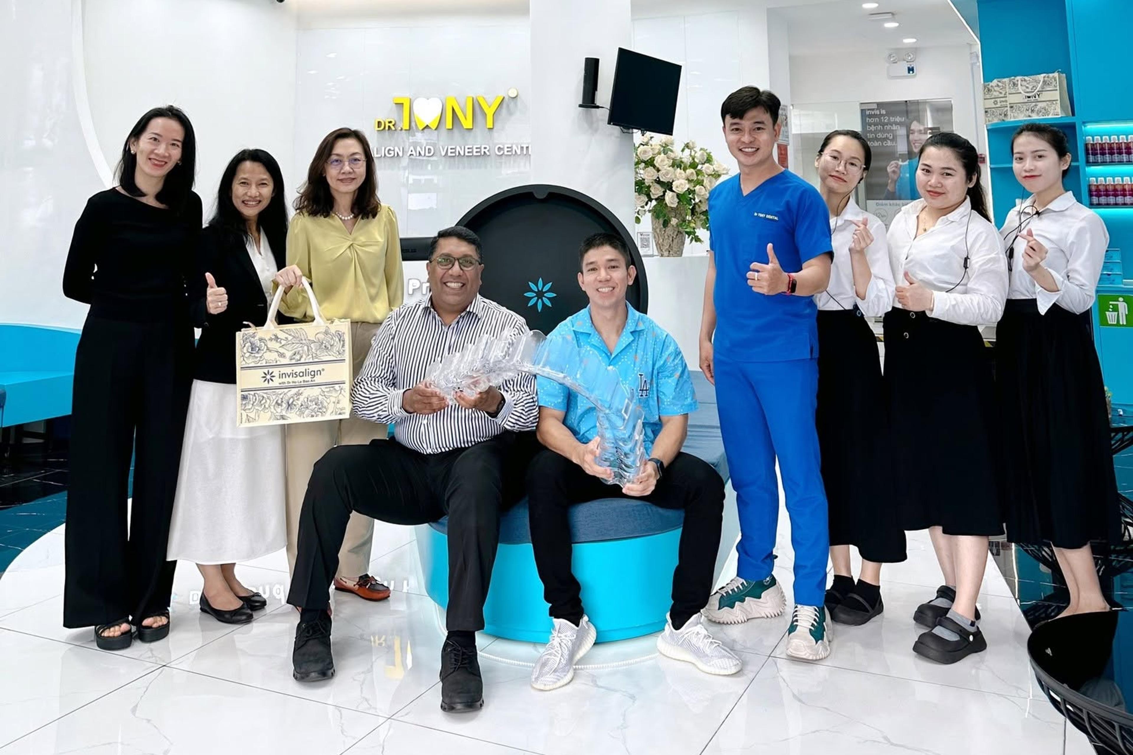 Đoàn lãnh đạo Align Technology chụp ảnh cùng đội ngũ Dr Tony Dental tại sảnh phòng khám; hai khách mời ngồi phía trước, một người cầm túi quà và một người cầm kỷ niệm chương pha lê.