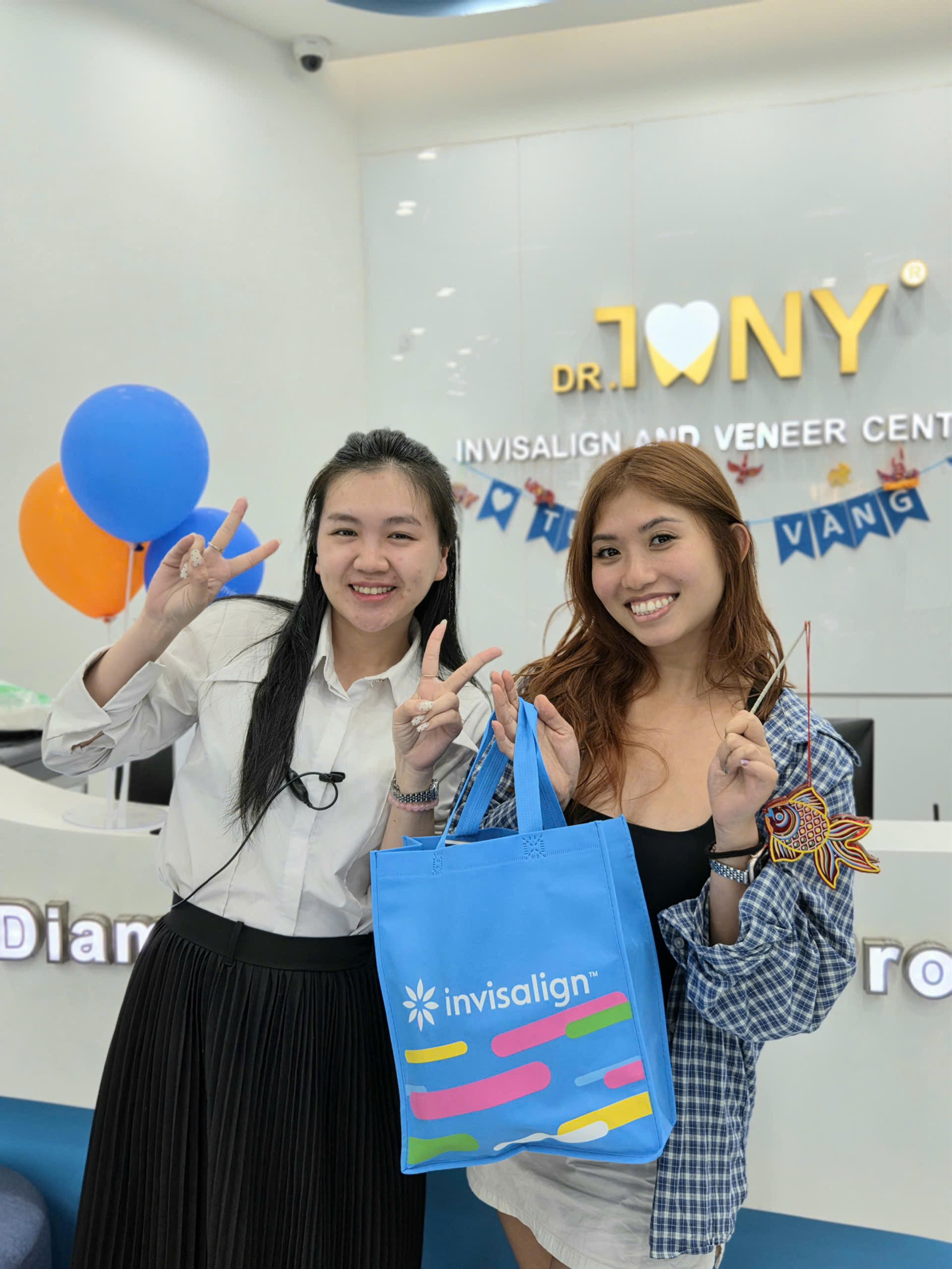 Hai người cười, giơ tay chữ V ở quầy lễ tân Dr Tony Dental; một người cầm túi vải xanh in chữ "invisalign", người kia cầm đèn lồng giấy hình cá chép. Phía sau là bảng hiệu "DR. TONY — INVISALIGN AND VENEER CENTER", dây cờ trang trí và chùm bóng bay xanh dương — cam.