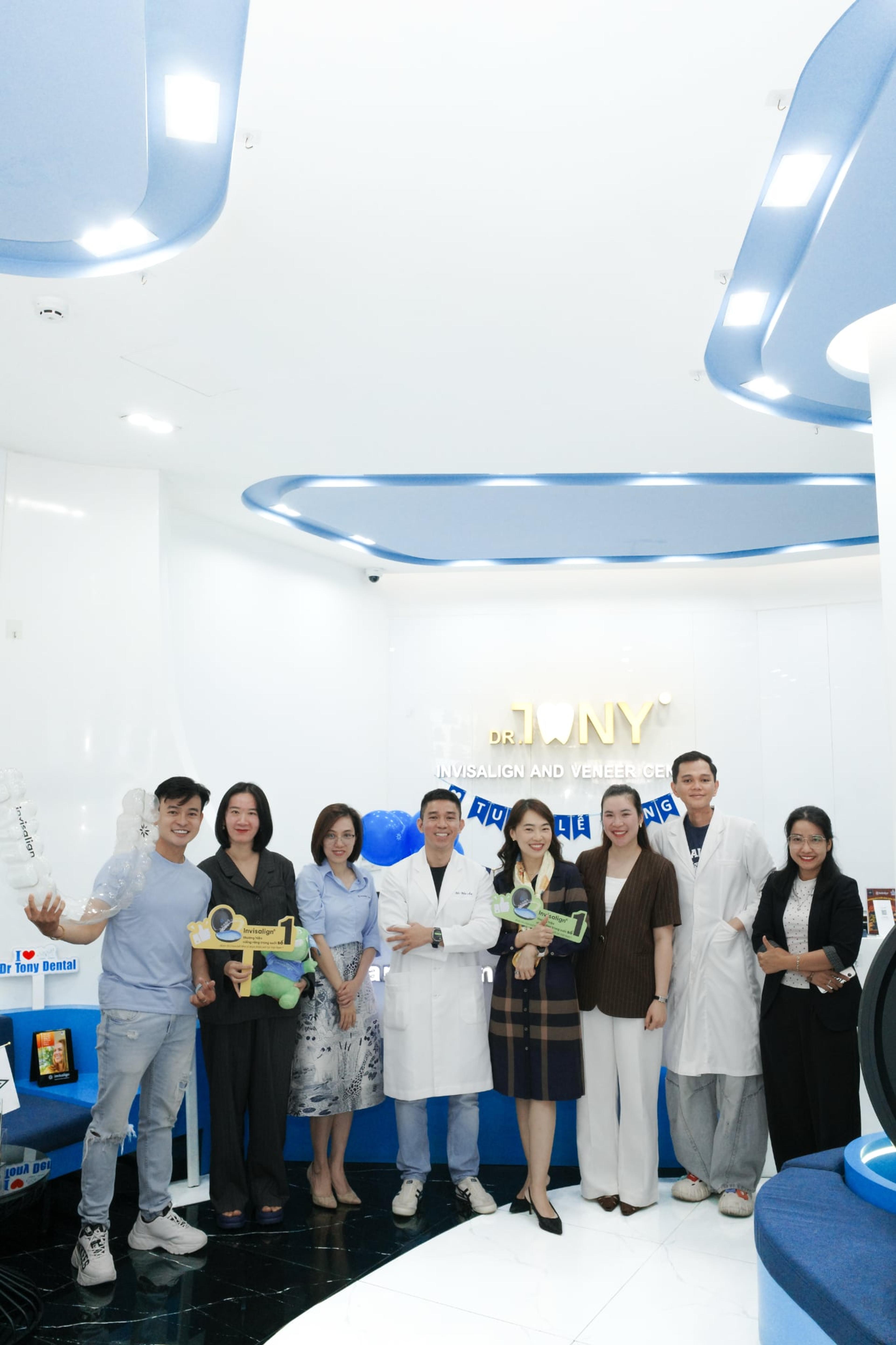 Cùng nhóm chín người ở sảnh phòng khám, chụp khung dọc lấy trọn trần đèn hắt xanh và bảng hiệu vàng “DR. TONY — INVISALIGN AND VENEER CENTER” phía sau.