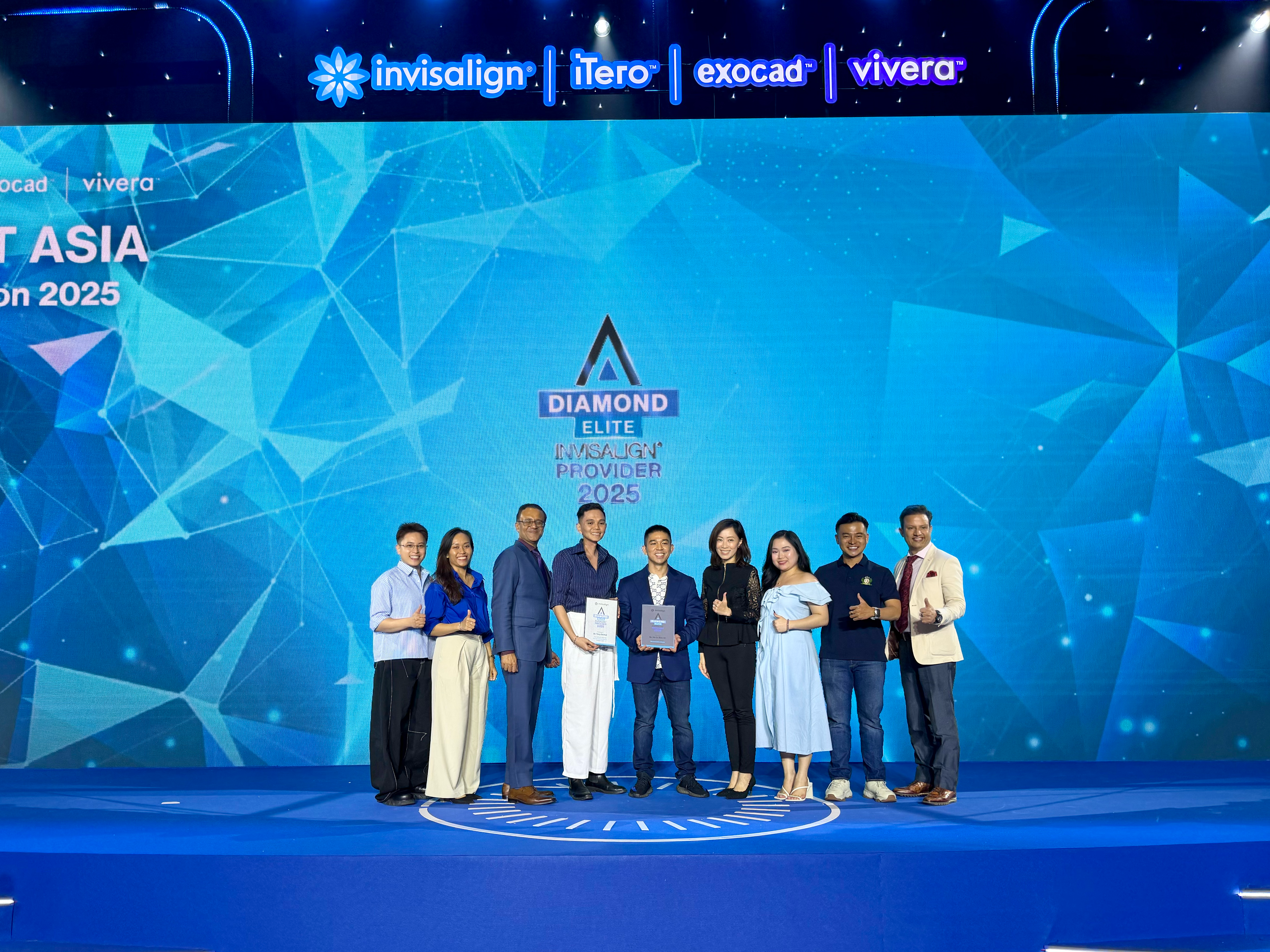Chín người đứng thành hàng trên sân khấu, màn hình phía sau ghi “Diamond Elite Invisalign Provider 2025”; hai người ở giữa cầm bảng chứng nhận.