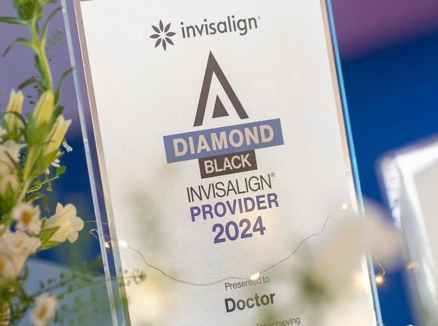 Cận cảnh bảng chứng nhận pha lê ghi “invisalign — DIAMOND BLACK — INVISALIGN PROVIDER 2024 — Presented to Doctor”, đặt cạnh bình hoa trắng.