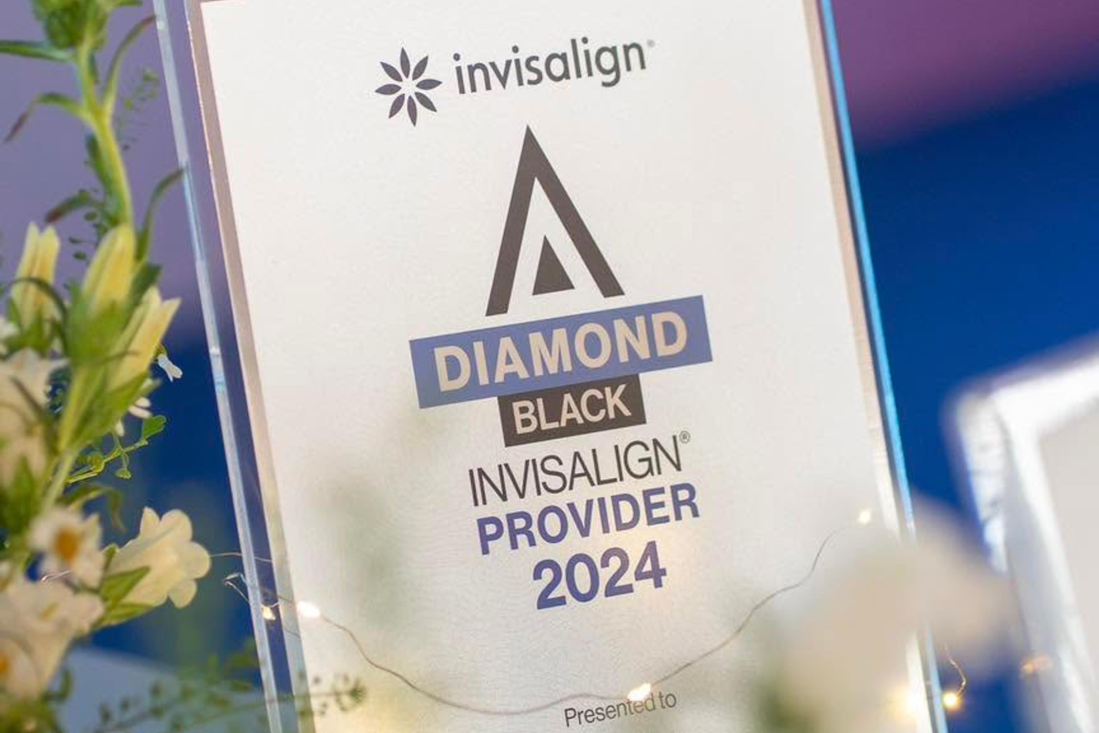 Cận cảnh bảng chứng nhận pha lê ghi “invisalign — DIAMOND BLACK — INVISALIGN PROVIDER 2024 — Presented to Doctor”, đặt cạnh bình hoa trắng.
