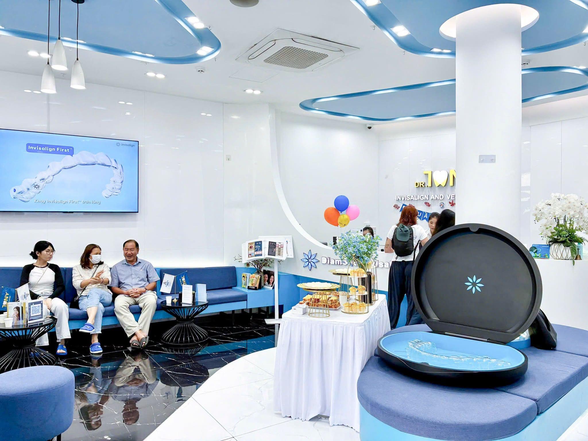 Khu chờ của phòng khám: ba khách ngồi trên ghế sofa xanh dưới màn hình giới thiệu khay Invisalign First; bên phải là mô hình hộp đựng khay Invisalign cỡ lớn đang mở nắp và bàn tiệc nhẹ, phía xa là quầy lễ tân với bảng hiệu Dr. Tony.