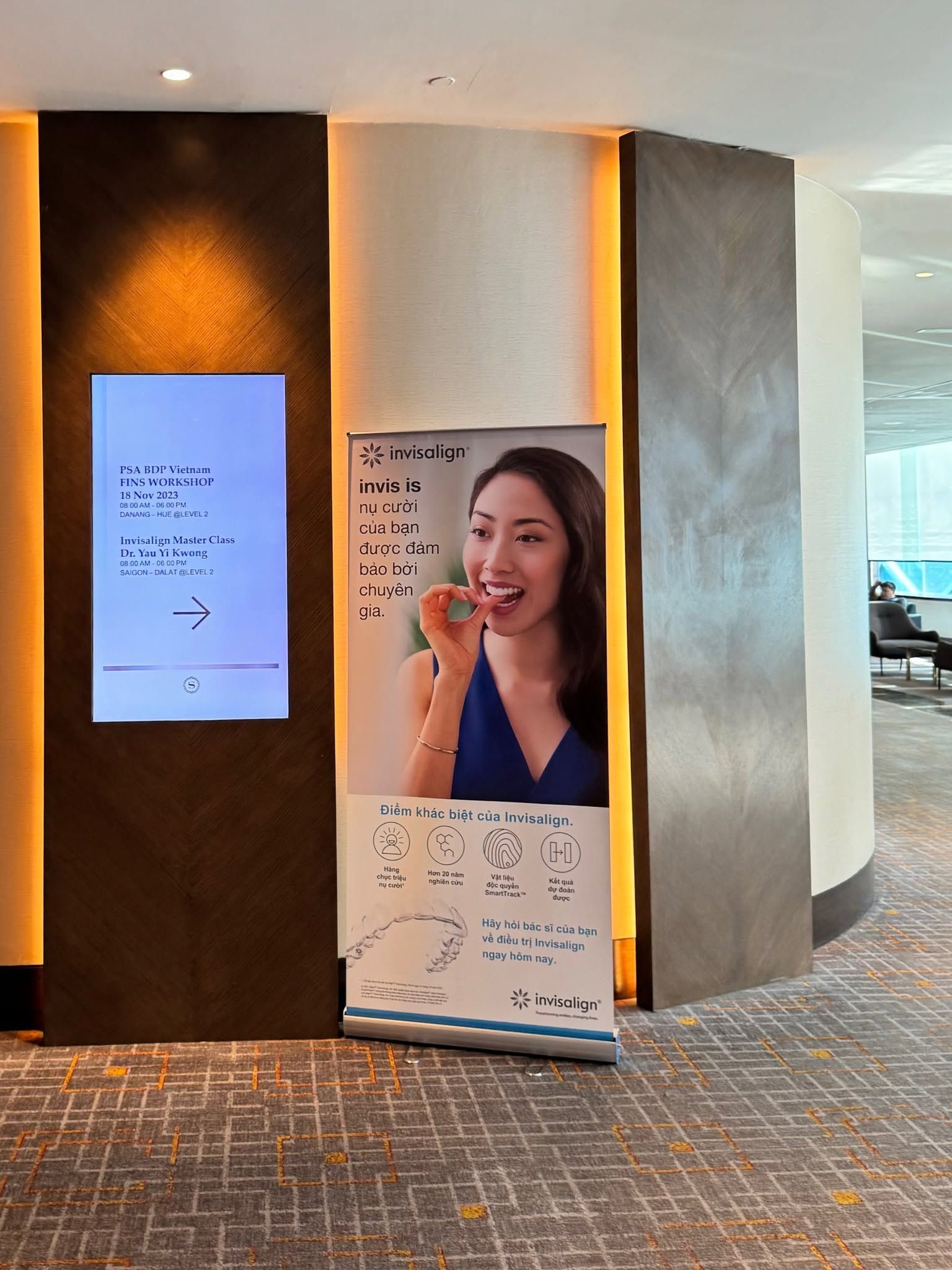 Bảng chỉ dẫn điện tử tại khách sạn ghi lịch chương trình “Invisalign Master Class – Dr. Yau Yi Kwong” ngày 18/11/2023, cạnh một standee Invisalign đặt trước lối vào phòng hội thảo.