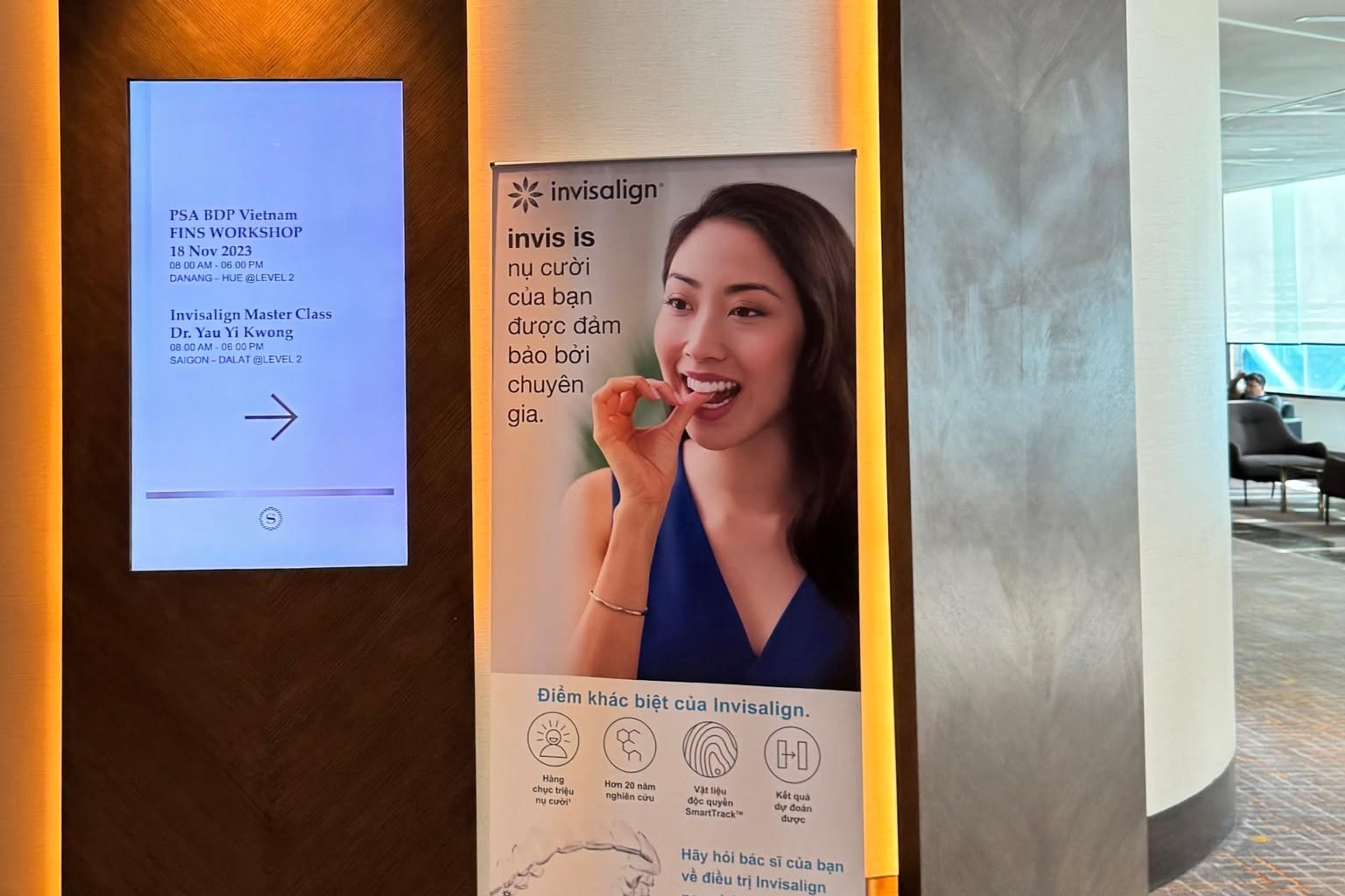 Bảng chỉ dẫn điện tử tại khách sạn ghi lịch chương trình “Invisalign Master Class – Dr. Yau Yi Kwong” ngày 18/11/2023, cạnh một standee Invisalign đặt trước lối vào phòng hội thảo.