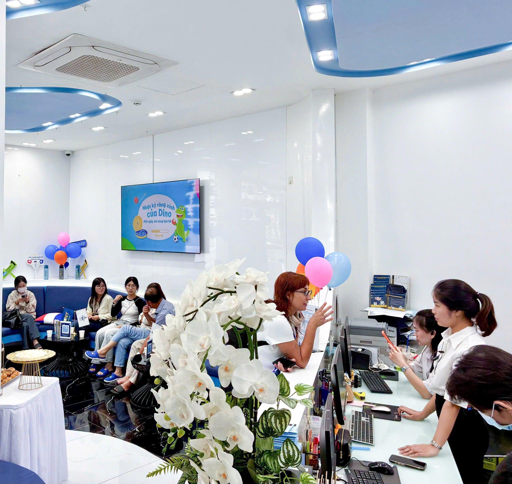 Quầy lễ tân Dr Tony Dental: một khách trò chuyện với nhân viên lễ tân bên bình lan hồ điệp trắng; phía sau, khách ngồi chờ ở dãy ghế xanh dưới màn hình chiếu "Nhật ký răng xinh của Dino" và chùm bóng bay xanh, hồng, cam.