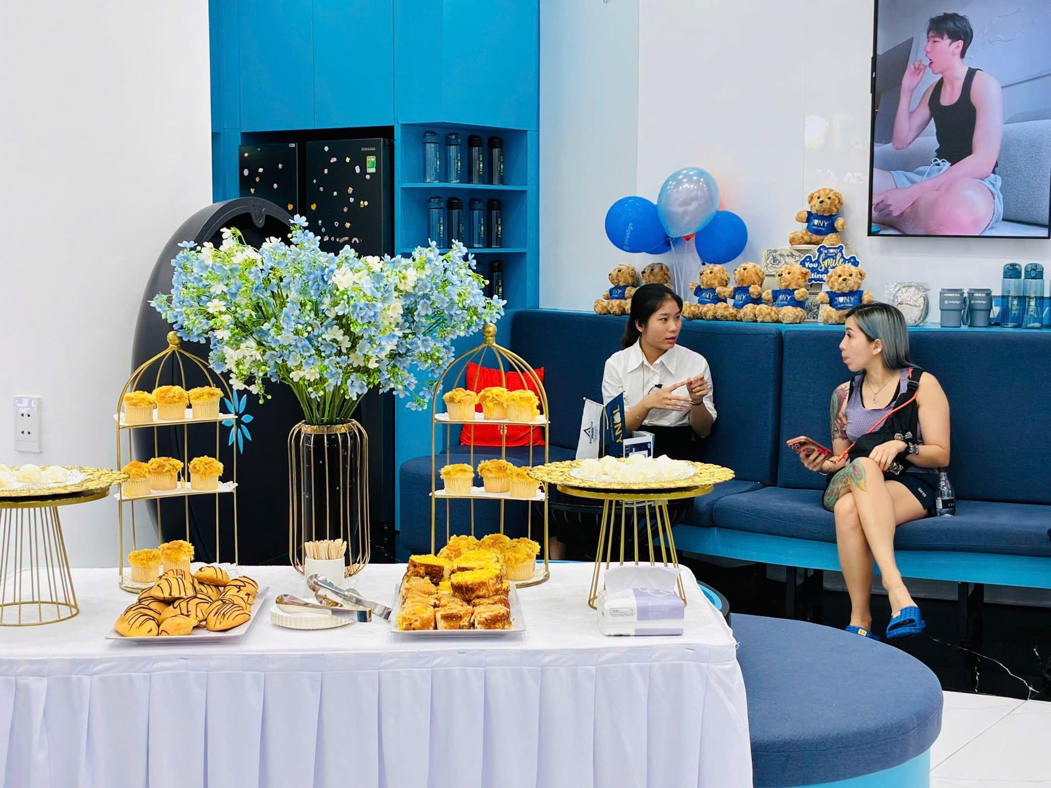 Một nhân viên phòng khám mặc sơ mi trắng trò chuyện với một khách trên ghế sofa xanh; phía trước là bàn tiệc nhẹ với bánh su, bánh tart và muffin trên các kệ vàng, cạnh bình hoa xanh trắng.