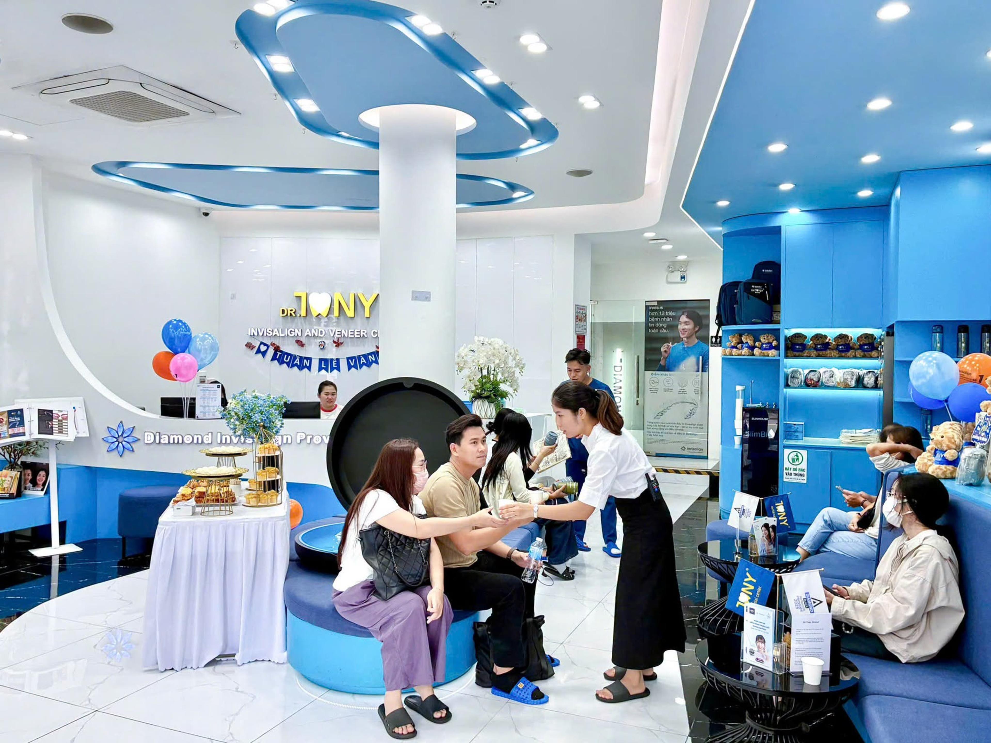 Sảnh chờ Dr Tony Dental trong ngày Open Day: một nhân viên mặc sơ mi trắng đón tiếp một cặp khách ngồi ở ghế tròn, bên trái là bàn tiệc nhẹ phủ khăn trắng; phía sau là bảng hiệu "DR. TONY — INVISALIGN AND VENEER CENTER", dòng chữ "Diamond Invisalign Provider" trên mặt quầy và kệ gấu bông nền xanh bên phải.