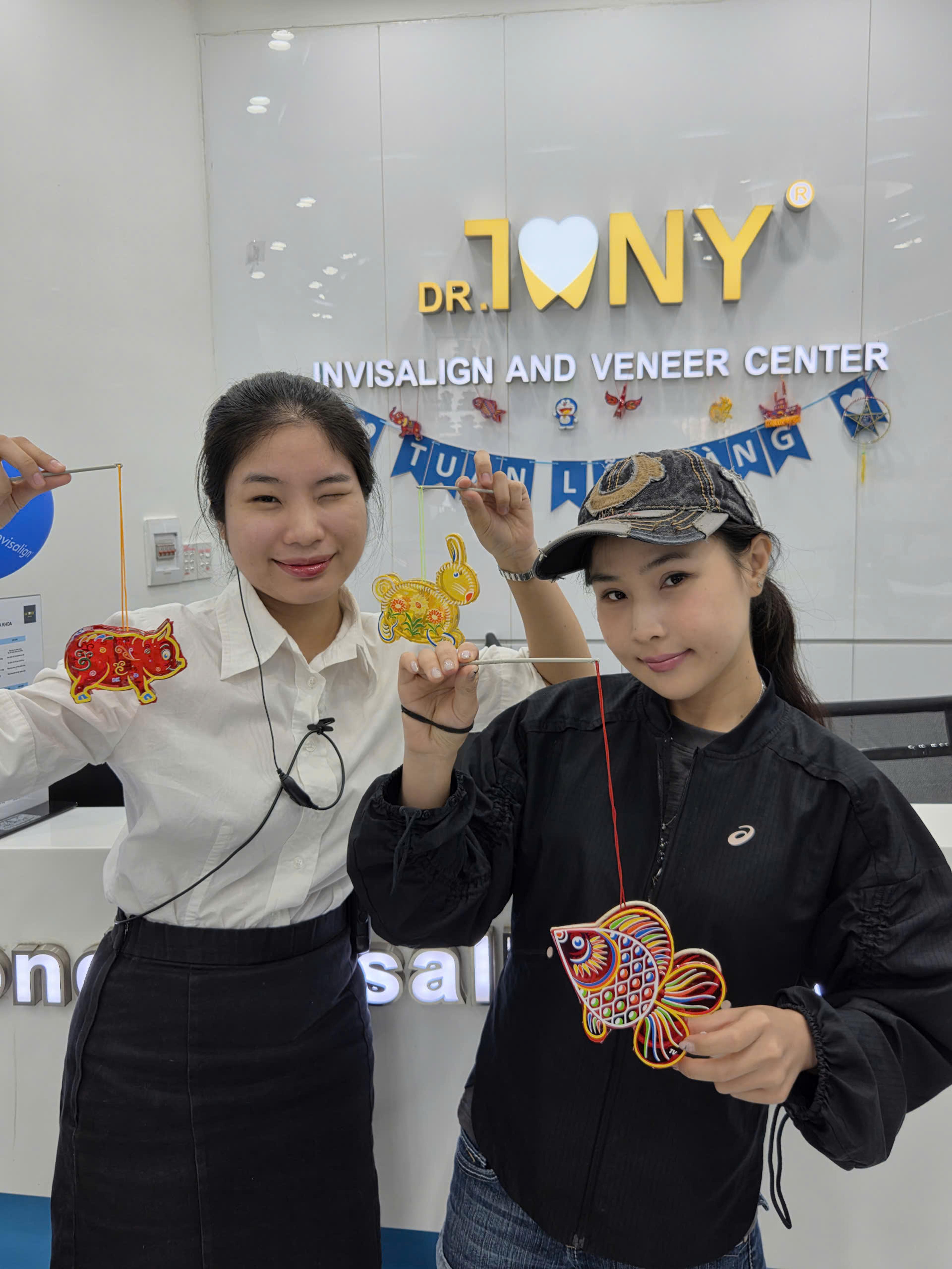 Hai người đứng cạnh quầy lễ tân Dr Tony Dental, cầm ba đèn lồng giấy thủ công hình con heo, con thỏ và con cá; phía sau là bảng hiệu "DR. TONY — INVISALIGN AND VENEER CENTER" và dây cờ trang trí treo các hình con giống.